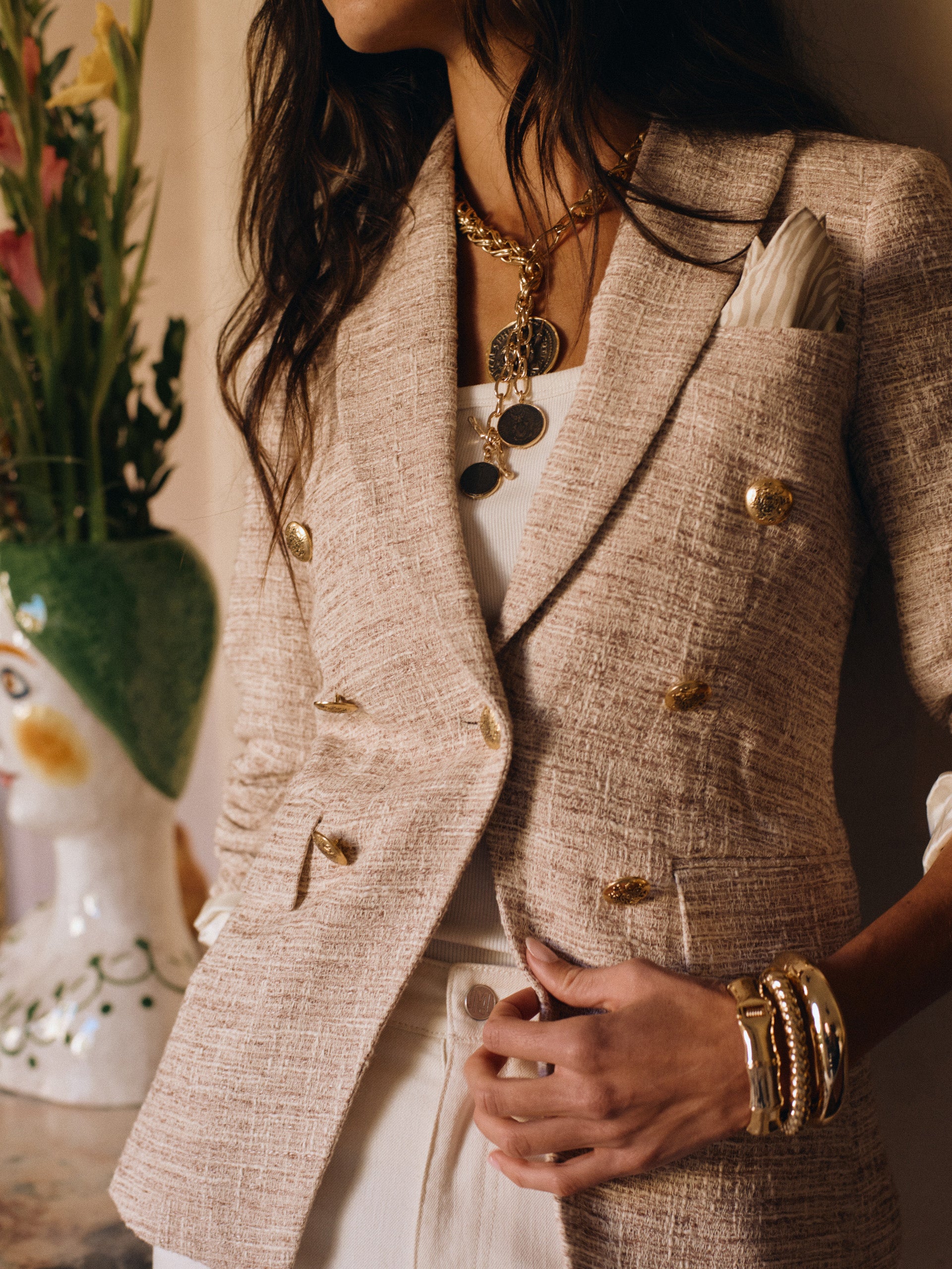 Woman wearing a beige textured Genevieve Tweed Blazer by J.McLaughlin with gold buttons, layered necklaces, and bracelets, styled with a white top and light pants.