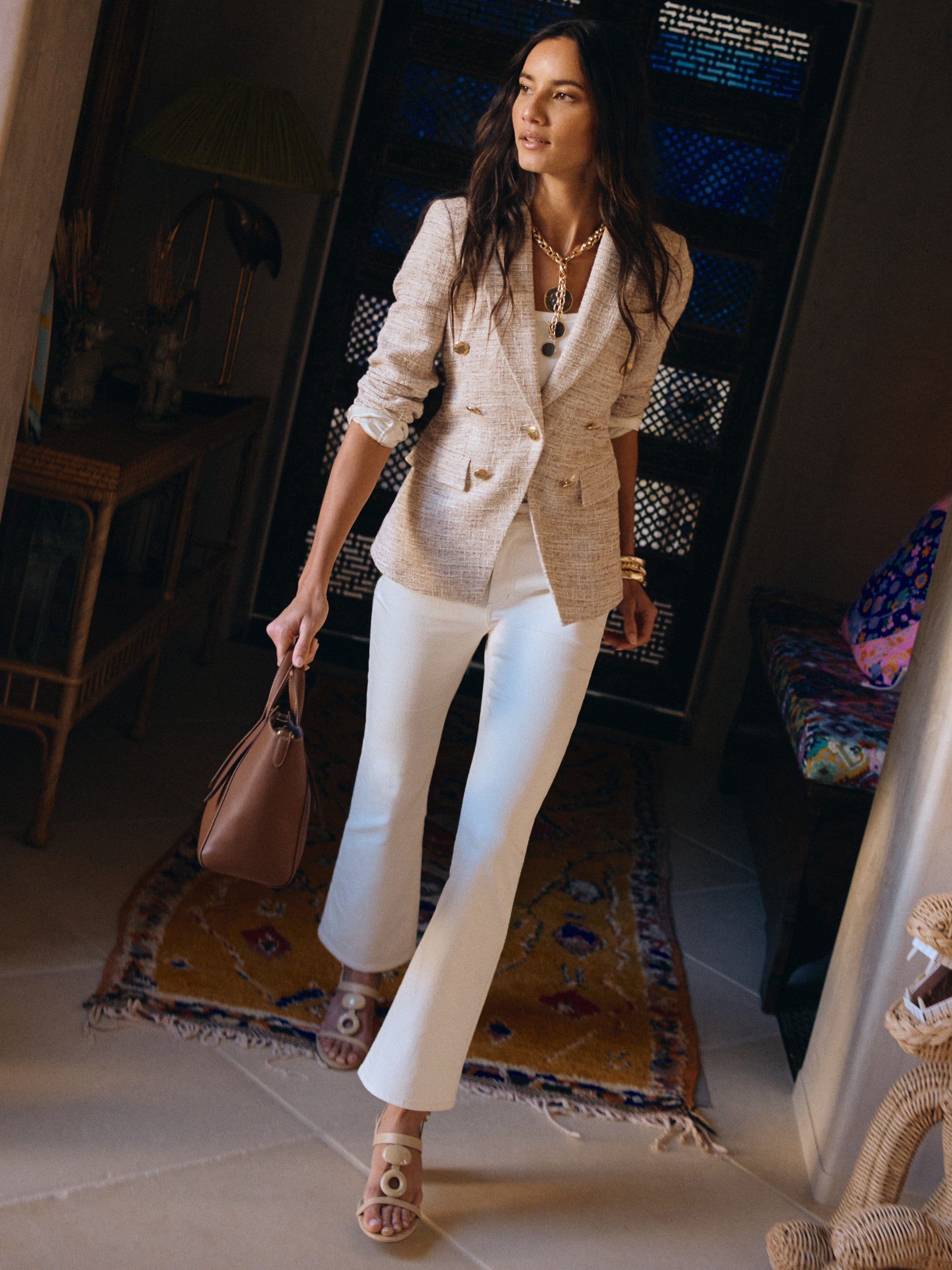 Woman wearing a stylish Genevieve Tweed Blazer by J.McLaughlin paired with white pants and sandals, carrying a brown handbag in an elegantly decorated room.