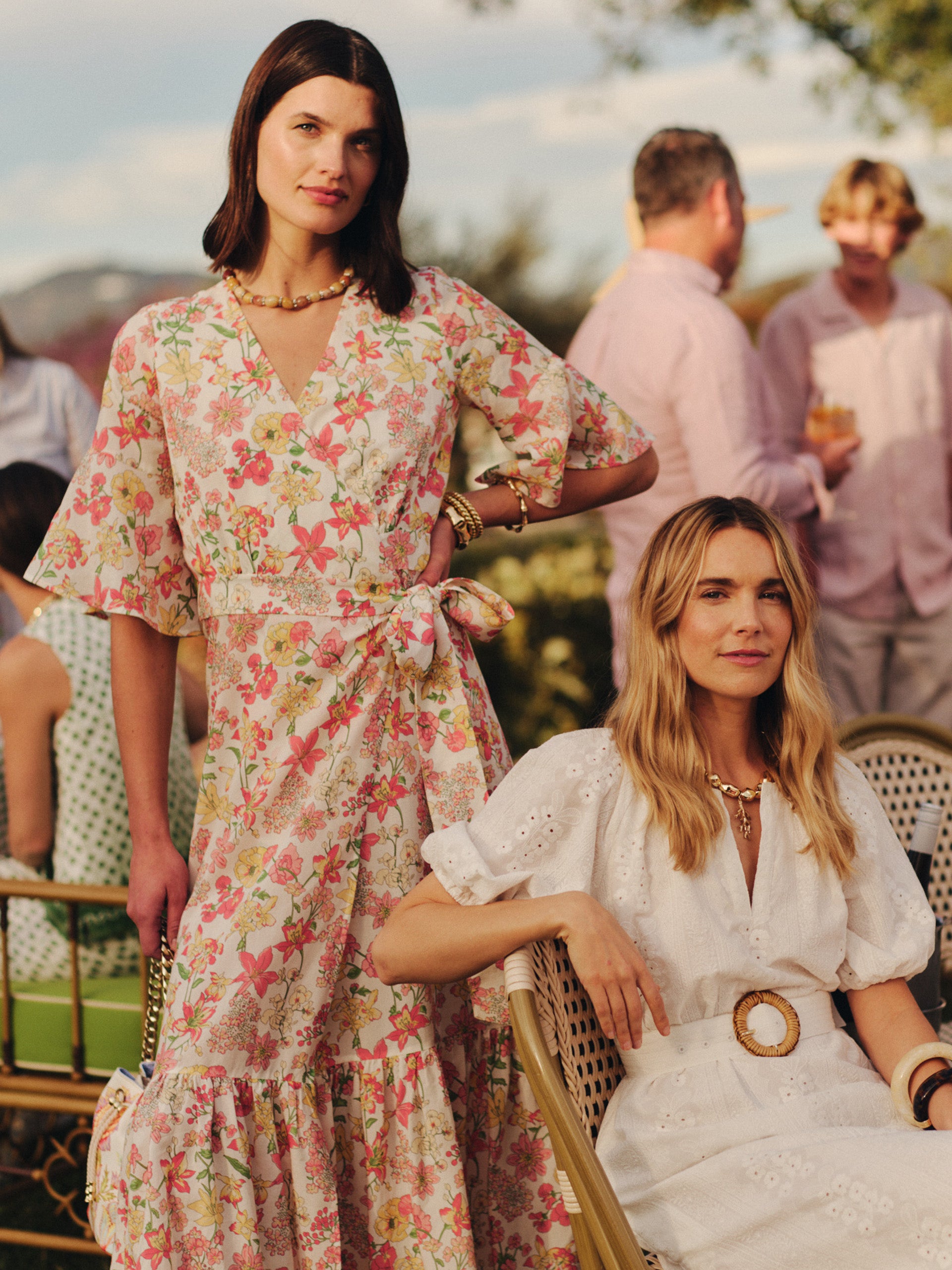Two women at an outdoor gathering, one standing and wearing the Emily Wrap Dress in Floral Villa by J.McLaughlin with pink and orange floral patterns, the other seated in a white dress with puff sleeves and a statement belt, both styled with gold jewelry.