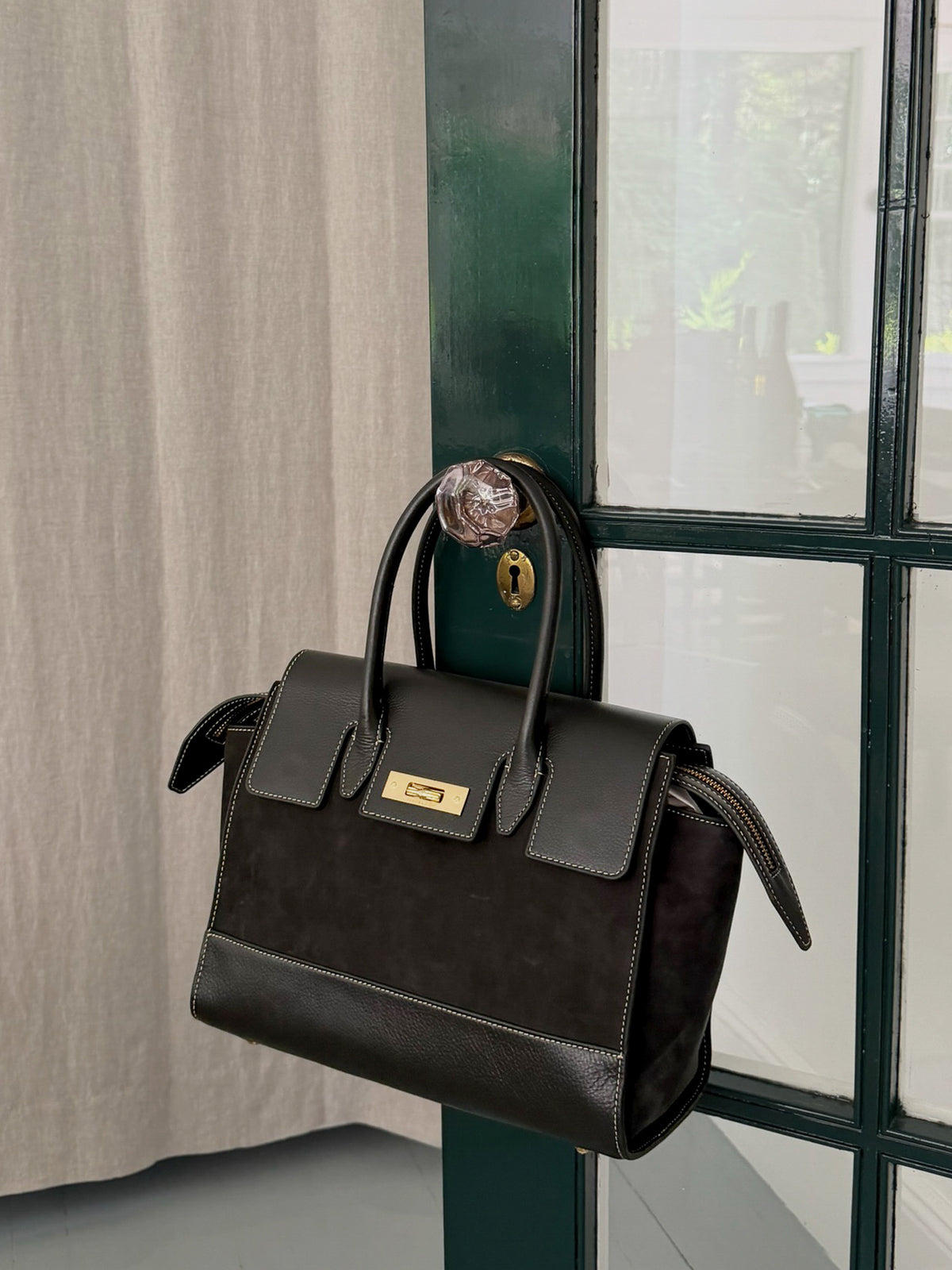 The Worth Leather Handbag by J.McLaughlin, a timeless black leather bag with gold-tone hardware, hanging on a dark green door handle, showcasing enduring style.