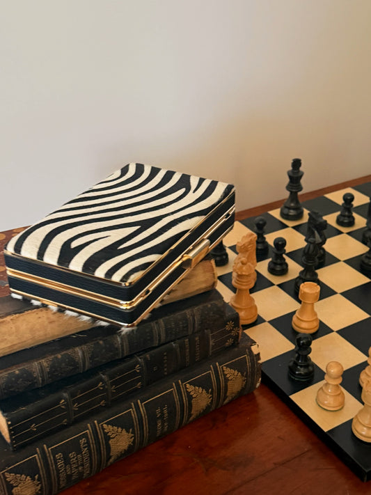 The Palm Pony Hair Clutch in Zebra Stripes by J.McLaughlin resting on vintage books next to a chessboard, showcasing an effortlessly stylish and timeless accessory.