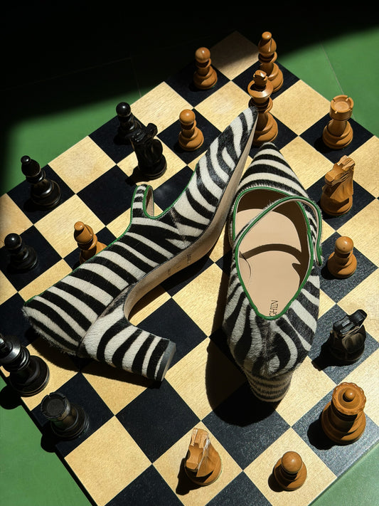 Katie Calf Hair Pumps in Zebra displayed on a chessboard, featuring a bold striped pattern and classic strap design by J.McLaughlin, blending timeless style with elegant footwear.