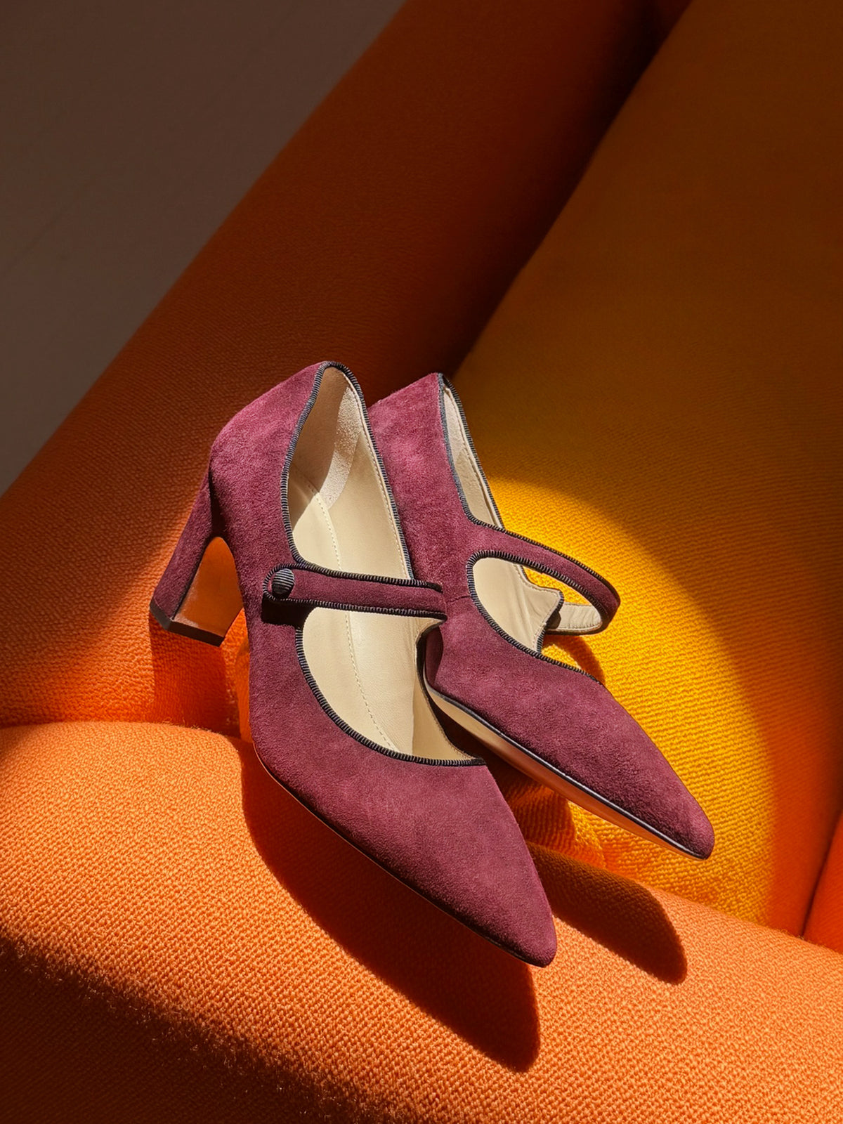 Katie Suede Pumps by J.McLaughlin in burgundy suede with pointed toes Mary Jane straps and slim heels styled on a warm orange surface