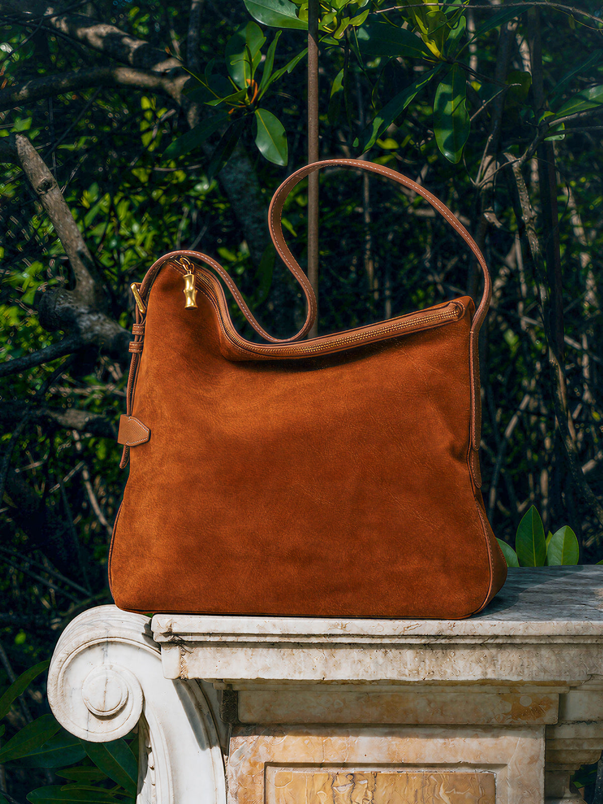 The Montecito Suede Hobo Bag by J.McLaughlin in rich brown suede with bamboo-inspired zipper pull resting on a stone ledge amidst lush greenery, showcasing timeless style.