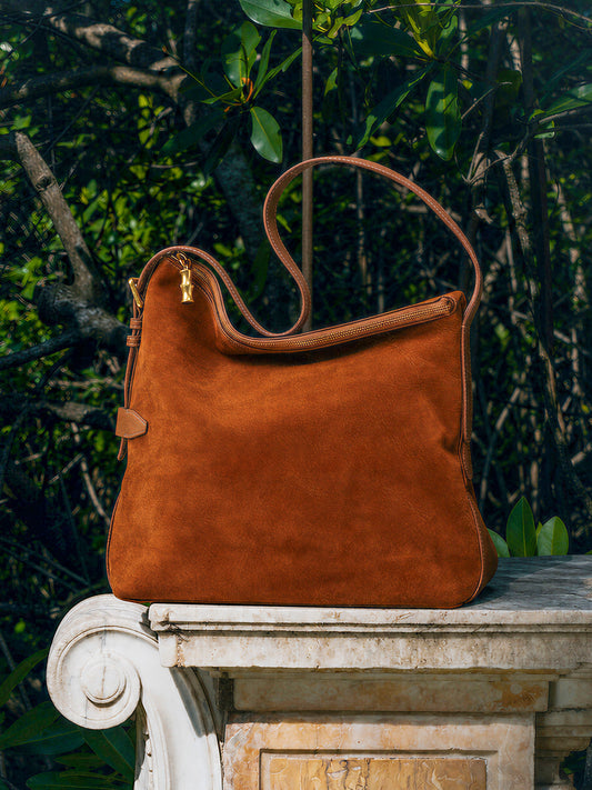 The Montecito Suede Hobo Bag by J.McLaughlin in rich brown suede with bamboo-inspired zipper pull resting on a stone ledge amidst lush greenery, showcasing timeless style.