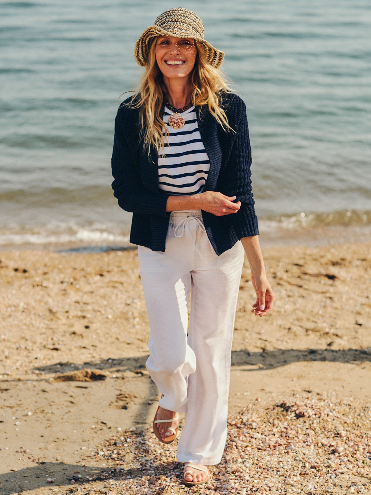 Woman walking on the beach wearing Irene Linen Pants by J.McLaughlin, paired with a navy cardigan and striped top, showcasing effortlessly stylish, enduring designs for a life well-lived.