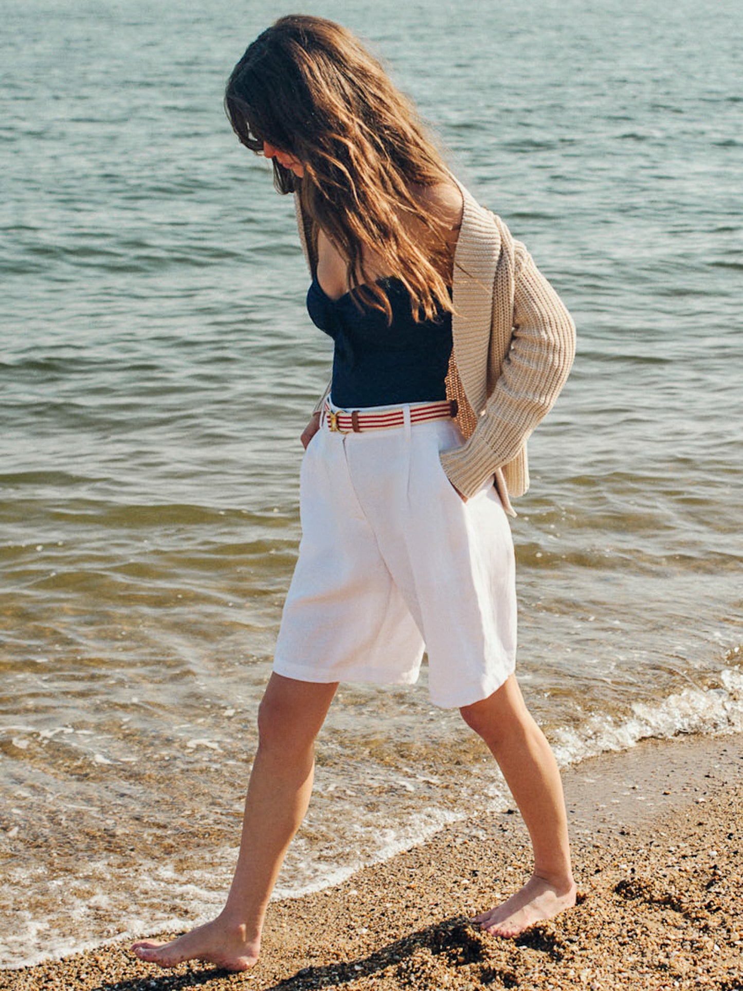 Woman walking barefoot on the beach wearing Cooper Linen Blend Shorts paired with a striped belt, a navy top, and a cozy oatmeal cardigan by J.McLaughlin, showcasing effortlessly stylish, enduring designs with innovative fabrics in a legacy of timeless fashion. A destination for defining style. Life, Well-Lived.