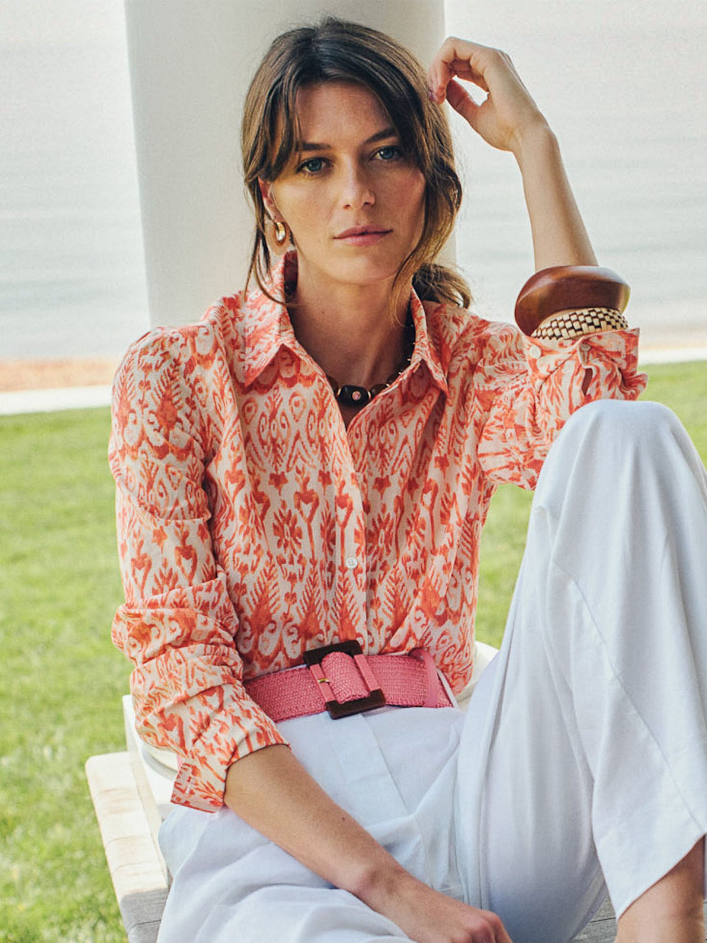 Woman wearing a patterned coral and cream blouse paired with white pants, accessorized with a pink Margot Grasscloth Belt by J.McLaughlin. Effortlessly stylish and enduring designs.