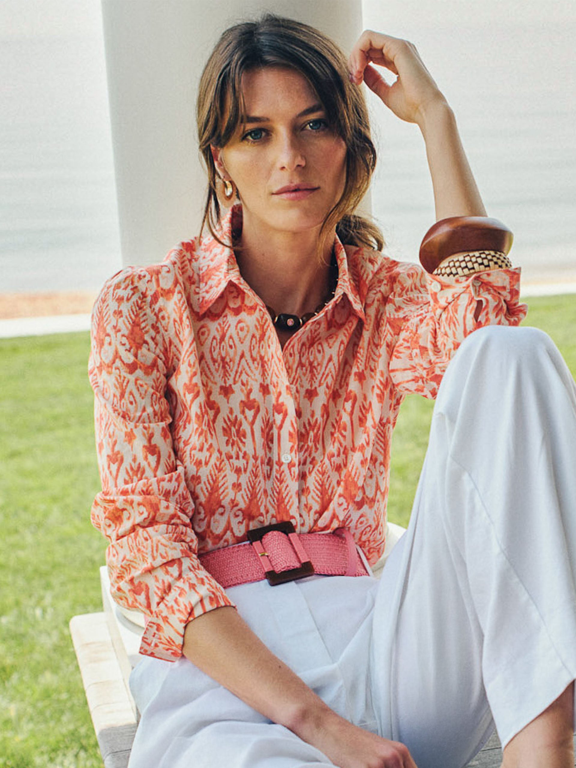 Woman wearing a patterned coral and cream blouse paired with white pants, accessorized with a pink Margot Grasscloth Belt by J.McLaughlin. Effortlessly stylish and enduring designs.