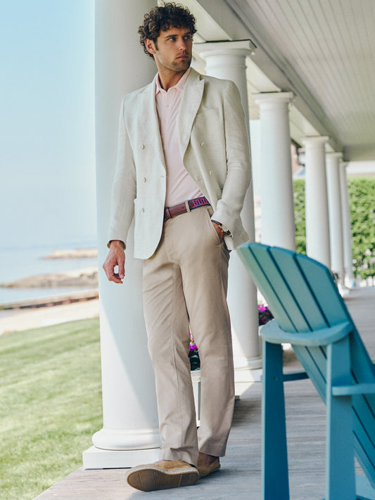 Man wearing Madison Pants by J.McLaughlin paired with a light blazer and pink shirt, standing on a porch overlooking a serene waterfront, showcasing effortlessly stylish, enduring designs.