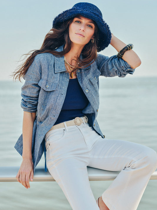 Woman wearing a blue Skye Linen Jacket by J.McLaughlin paired with white pants and a navy hat, showcasing effortlessly stylish and enduring designs with a legacy of timeless fashion.