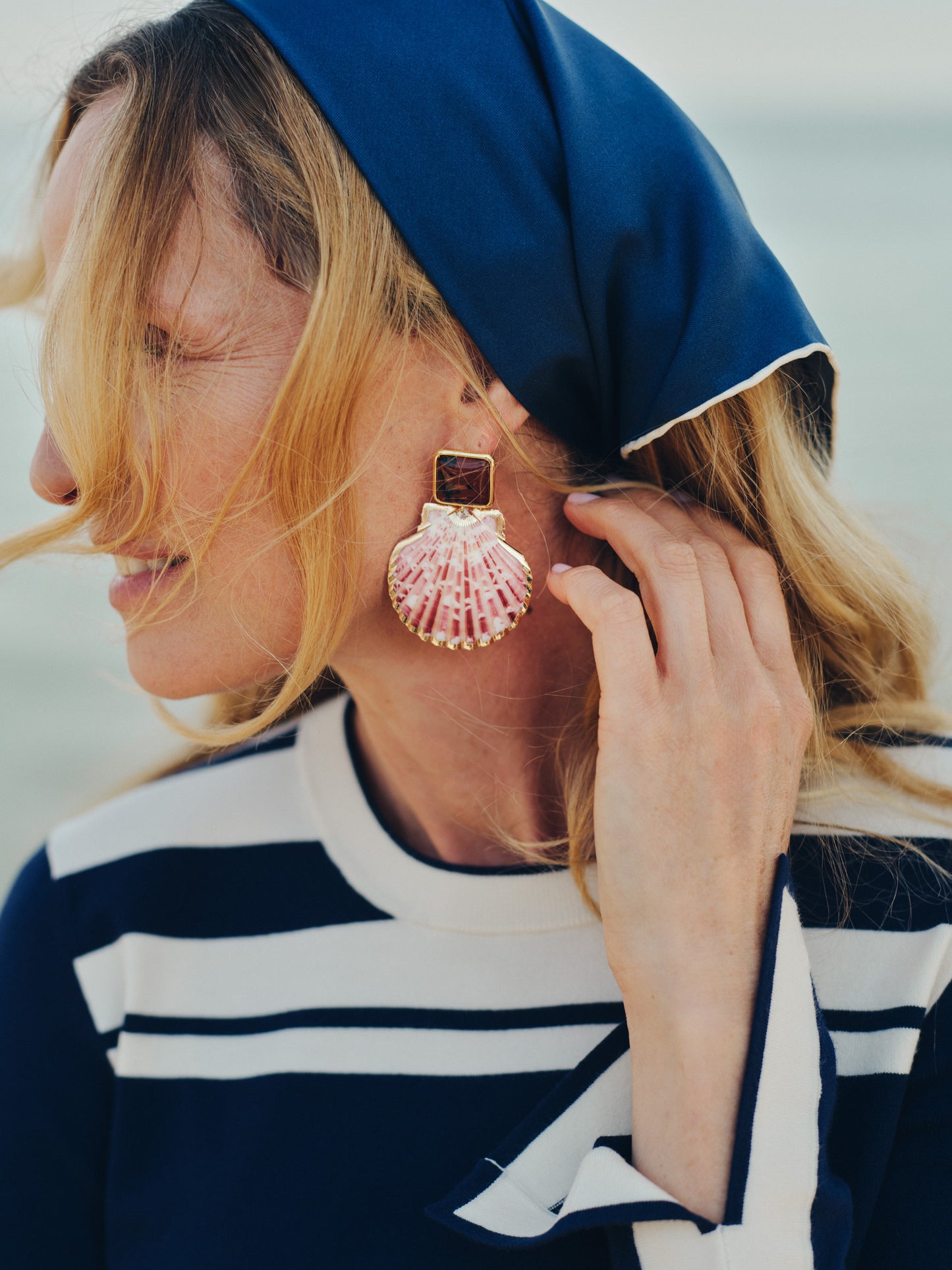 Close-up of a woman wearing J.McLaughlin Shell Earrings with a pink shell design, paired with a navy and white striped top and navy headscarf, embodying effortlessly stylish and enduring designs.