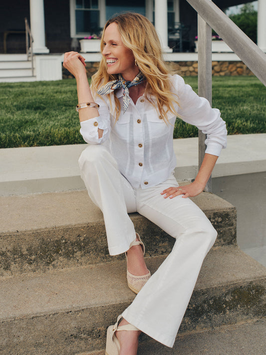 Woman sitting on concrete steps wearing a white Halo Linen Jacket by J.McLaughlin, paired with white pants and a patterned neck scarf, embodying effortlessly stylish, enduring designs and innovative fabrics that reflect a legacy of timeless fashion. A destination for defining style. Life, Well-Lived.