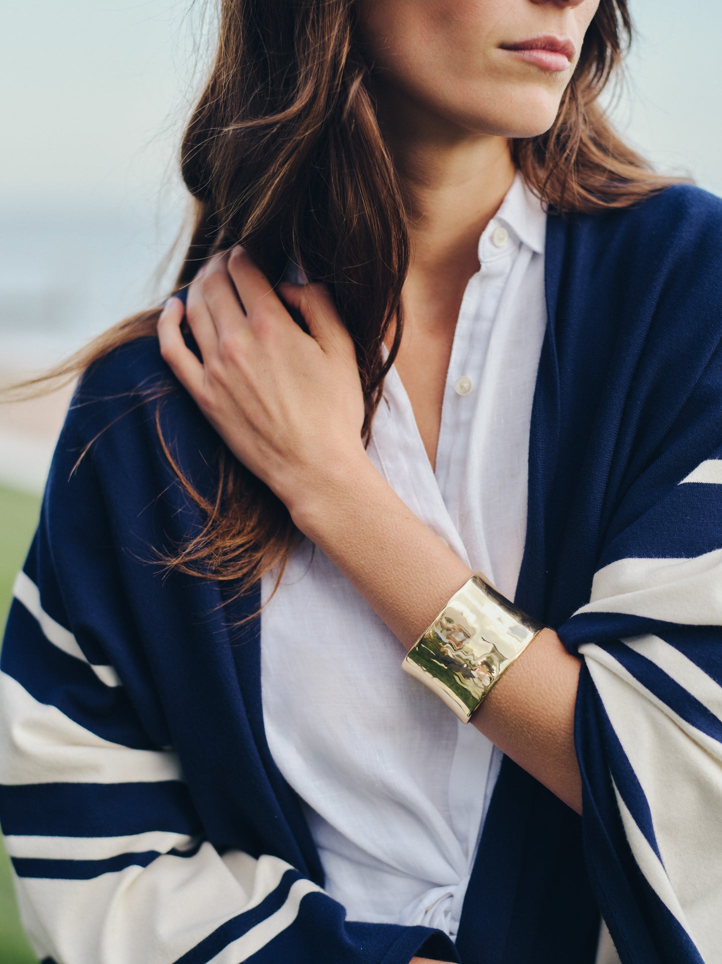 Woman wearing a Thick Gold Cuff bracelet by J.McLaughlin paired with a classic white shirt and navy striped wrap, embodying effortlessly stylish, enduring designs for a life well-lived.