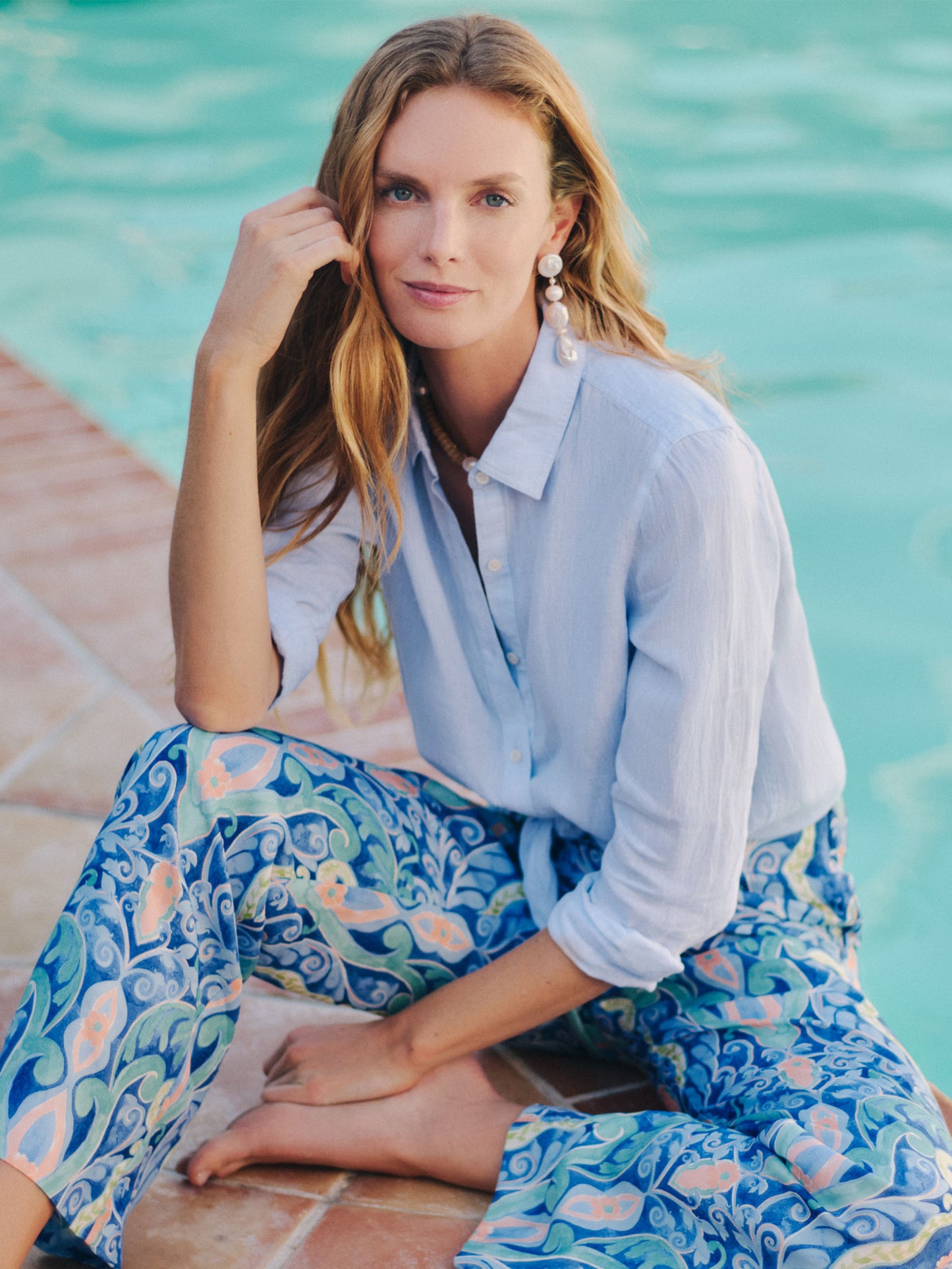 Woman wearing a light blue Britt Linen Shirt by J.McLaughlin, styled with patterned blue and coral pants, sitting relaxed by a pool, embodying effortlessly stylish, enduring designs, and innovative fabrics. A destination for defining style. Life, Well-Lived.