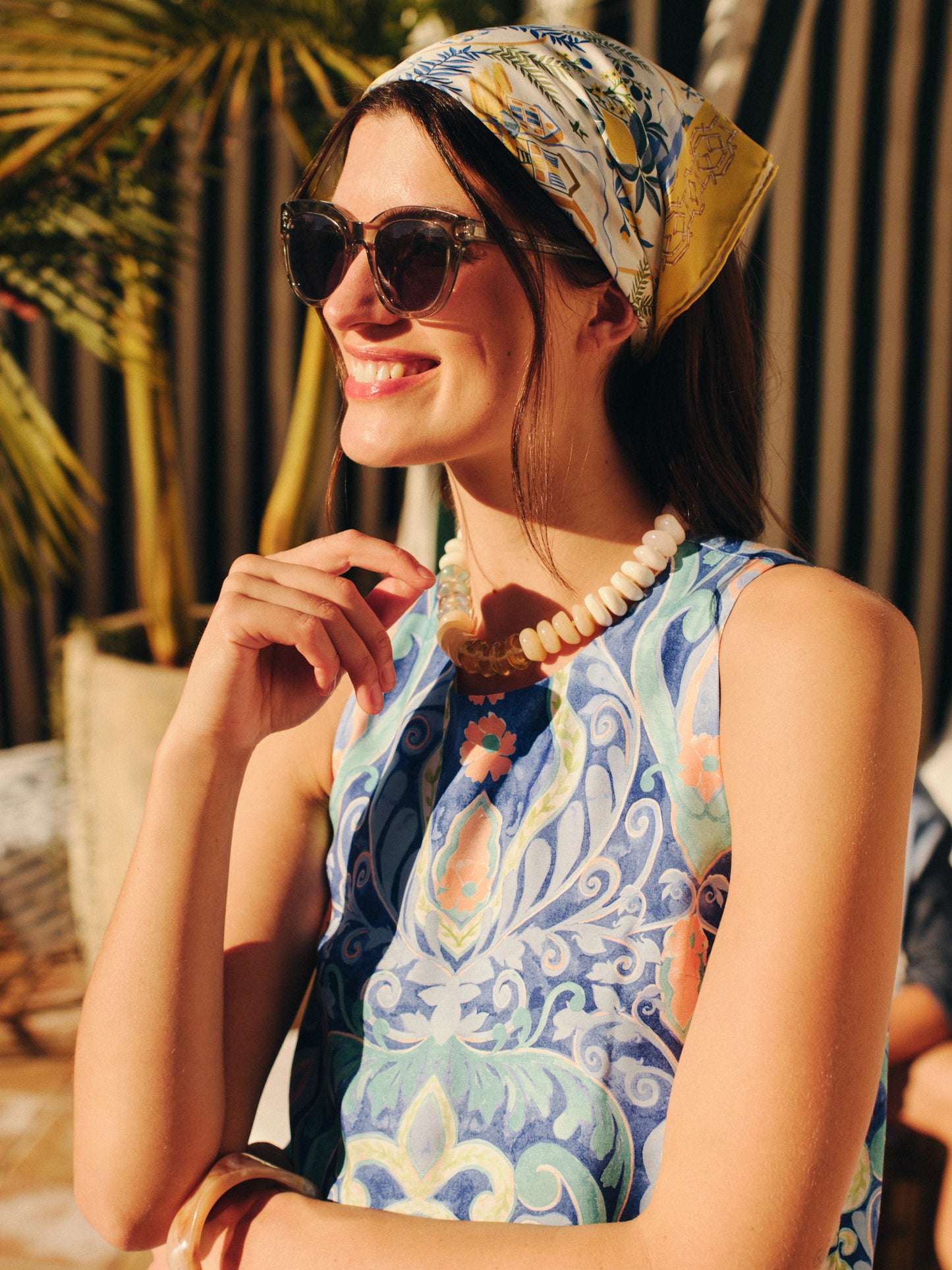 Woman wearing the Nikka Silk Scarf in Amalfi Coast Bamboo styled with sunglasses and a patterned sleeveless top, showcasing J.McLaughlin’s effortlessly stylish, enduring designs in a sunny outdoor setting.