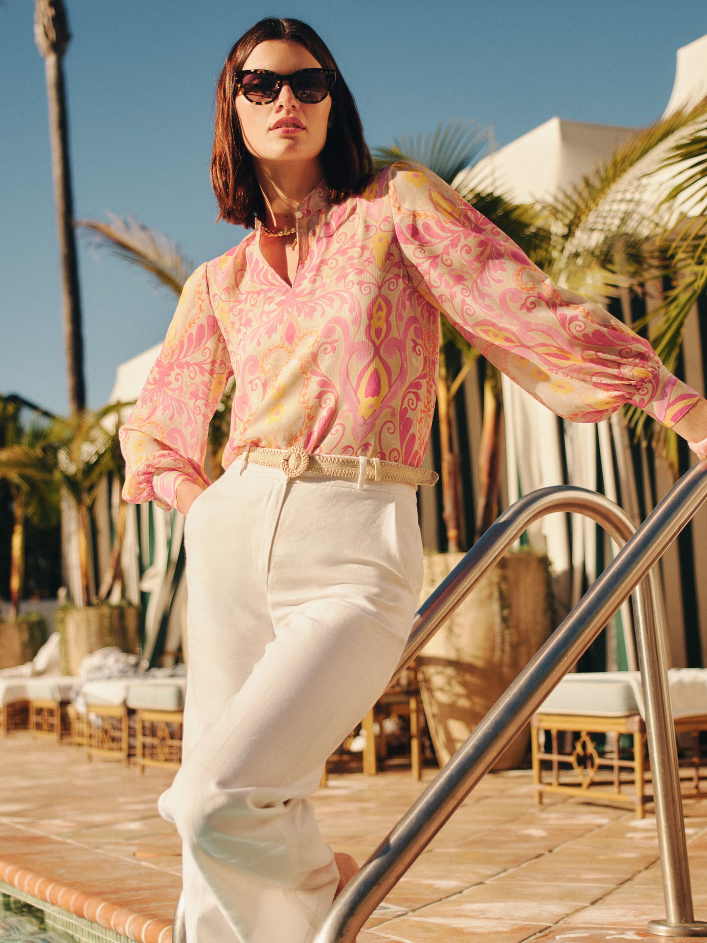 Woman wearing a Peyton Blouse in Mini Deco Bloom by J.McLaughlin in pink and yellow pattern, paired with cream pants, posing poolside by palm trees, showcasing effortlessly stylish, enduring designs and innovative fabrics for a legacy of timeless fashion. A destination for defining style. Life, Well-Lived.