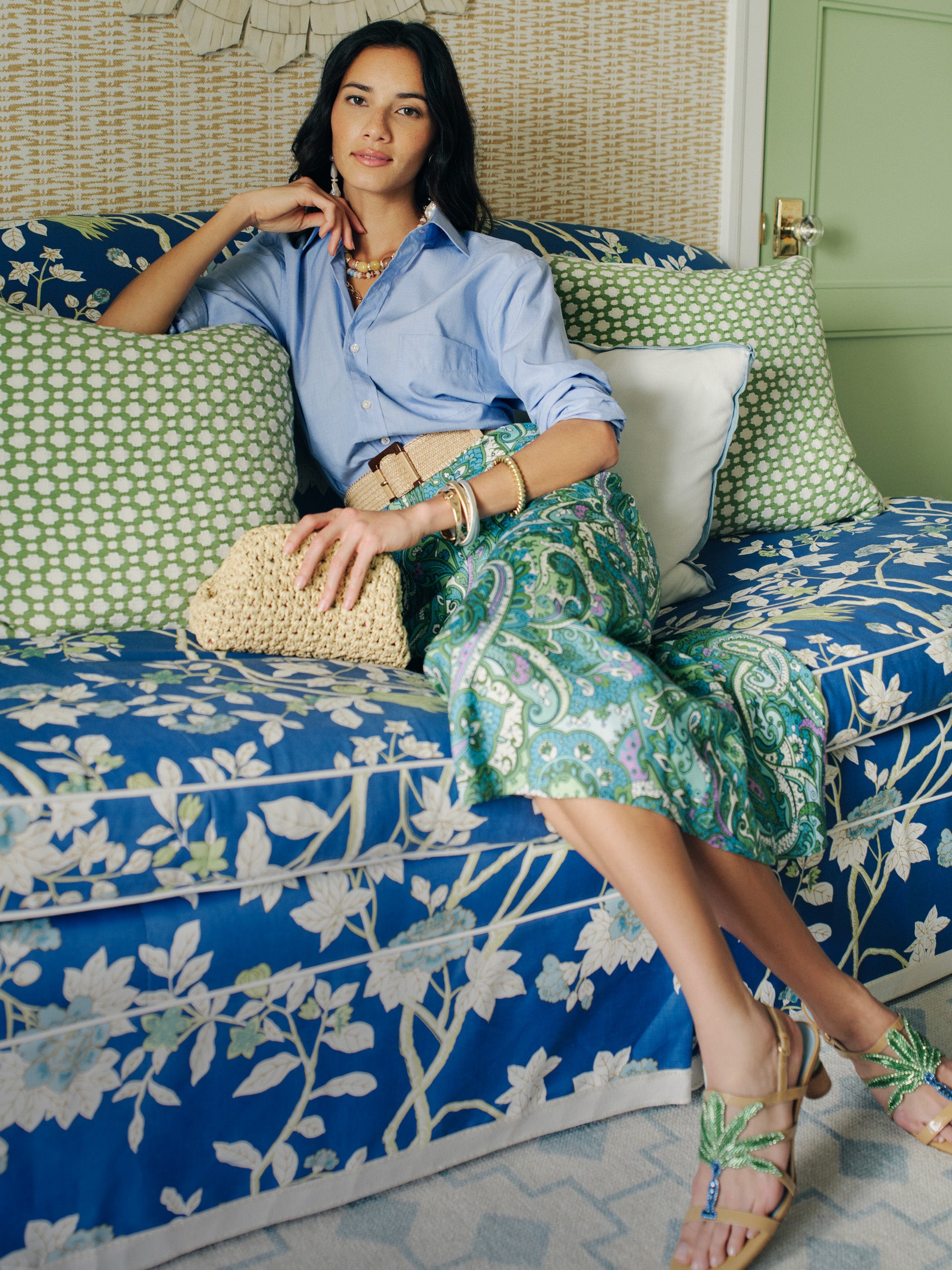 A stylish woman seated on a floral blue couch wearing the Adelyn Skirt in Paisley Reverie Bias by J.McLaughlin, paired with a light blue shirt and woven clutch, embodying effortlessly stylish, enduring designs in a cozy setting.