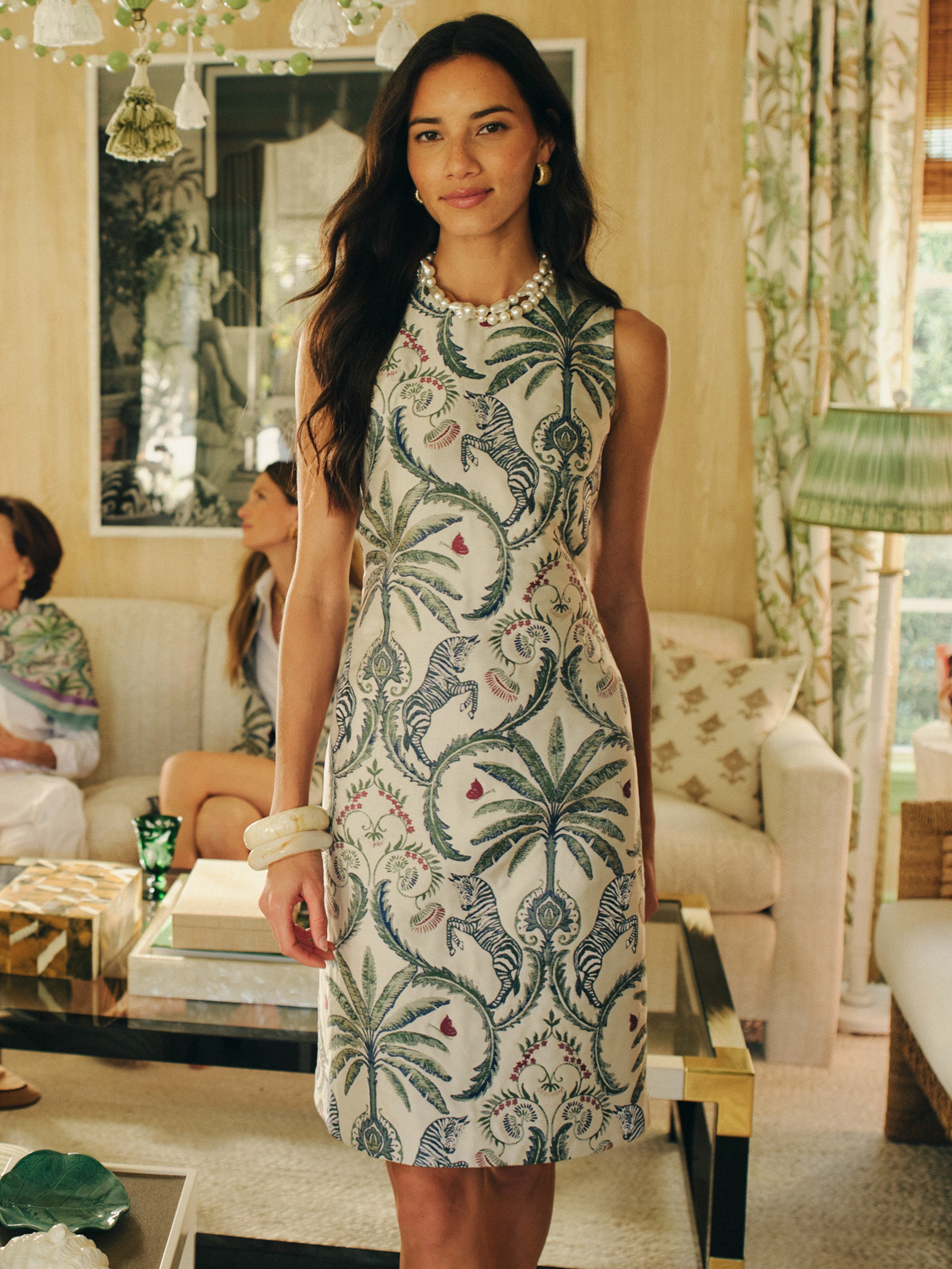 Woman wearing a sleeveless Kinsley Dress in Maxi Zebra Isle Jacquard by J.McLaughlin, featuring an elegant zebra and palm tree pattern, embodying effortlessly stylish, enduring designs with innovative fabrics in a classic living room setting.