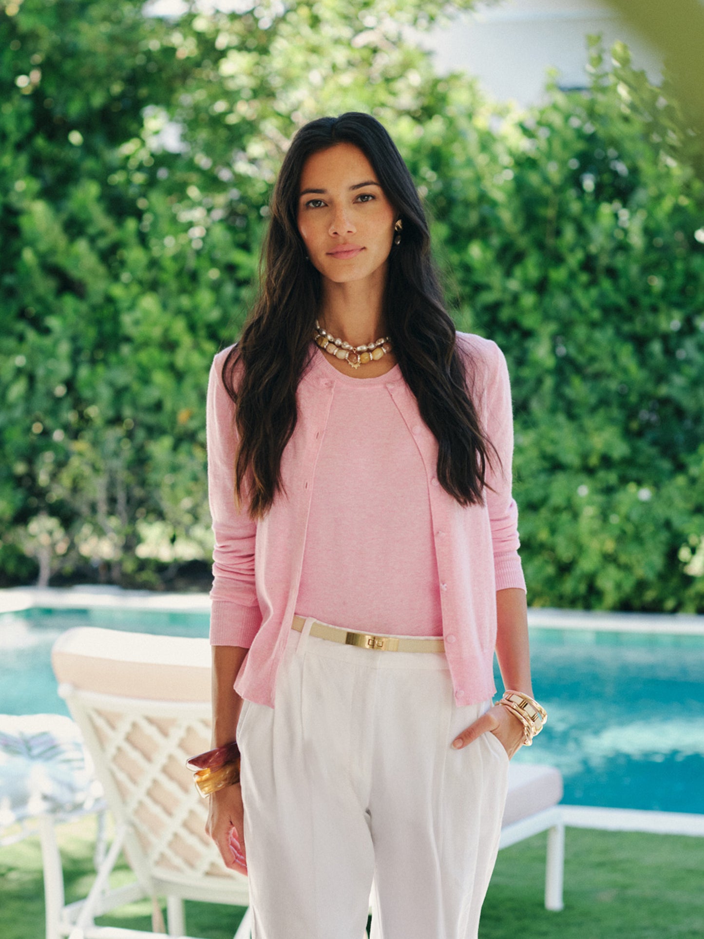 Woman wearing a pink Opal Sweater Tank by J.McLaughlin paired with white pants and gold accessories, standing outdoors near a pool surrounded by green foliage, effortlessly stylish and enduring designs.