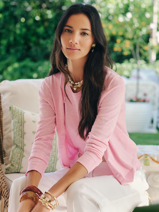 Woman seated outdoors wearing a pink Pearl Cardigan from J.McLaughlin, styled with white pants and gold accessories, showcasing effortlessly stylish, enduring designs and innovative fabrics.