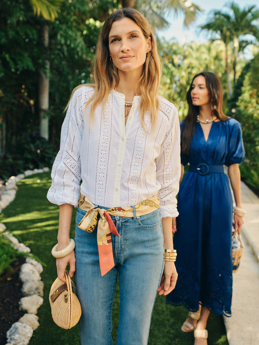 A woman wearing the Hazel Blouse in Eyelet by J.McLaughlin pairs it with jeans and a silk scarf belt, showcasing effortlessly stylish, enduring designs in a garden setting with another woman in a blue dress in the background.