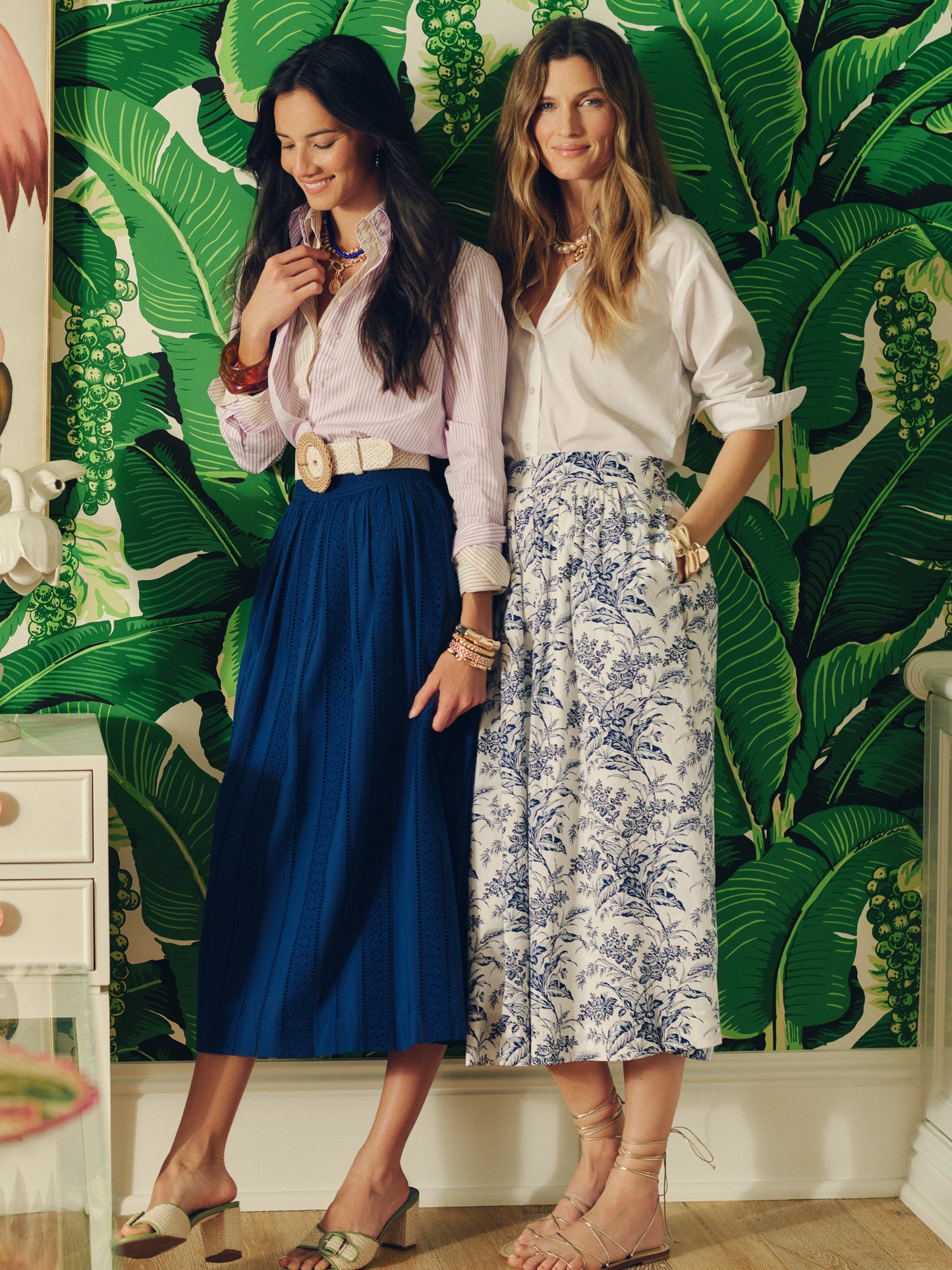 Two women styled in timeless elegance, one wearing the Winnie Skirt in Eyelet by J.McLaughlin in navy, paired with a light blouse and accessories, showcasing effortlessly stylish, enduring designs in a vibrant setting, embodying innovative fabrics and a legacy of timeless fashion at a destination for defining style. Life, Well-Lived.