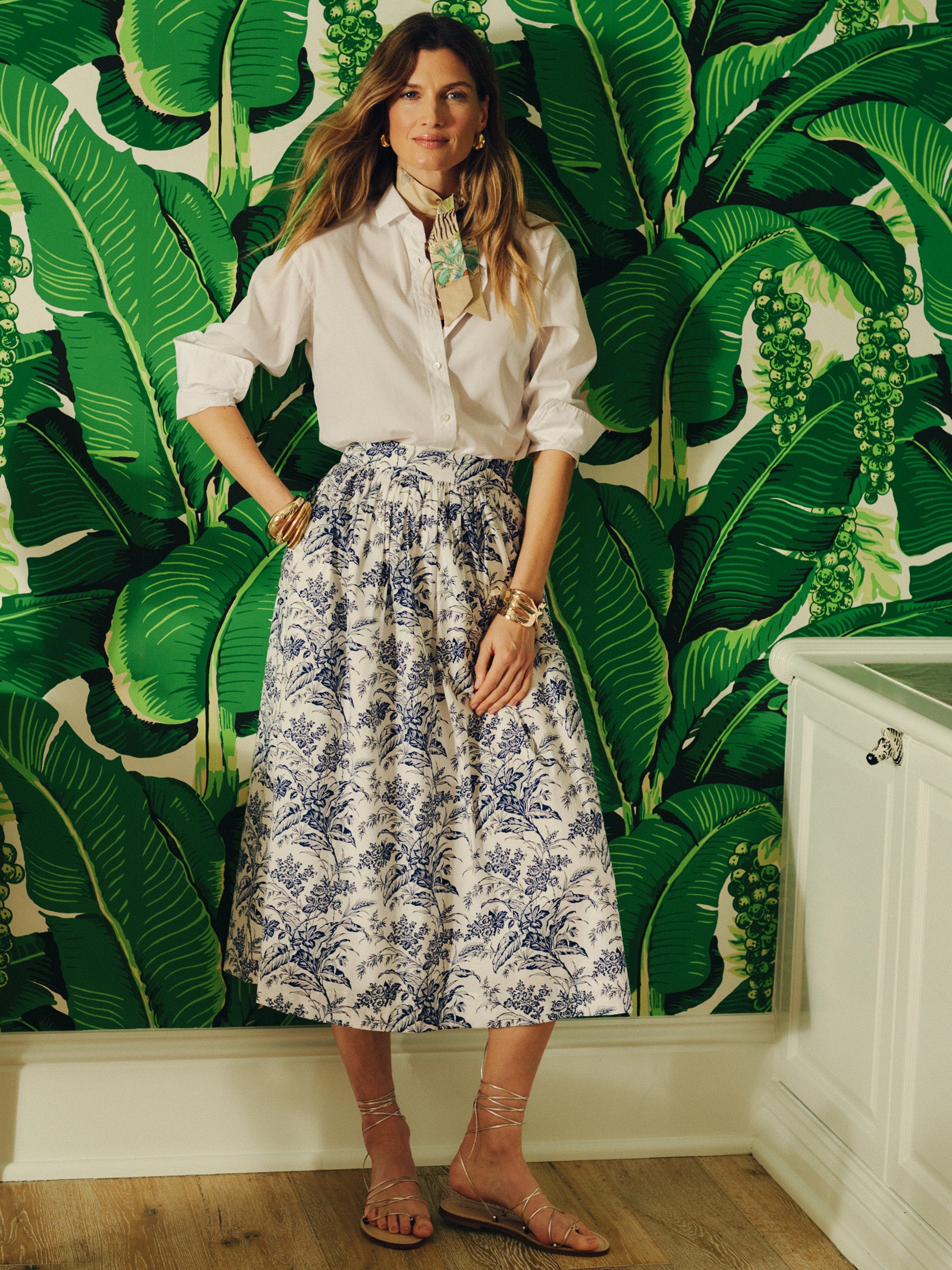 Woman wearing a white blouse and the Winnie Skirt in Giardino Toile by J.McLaughlin, standing against a vibrant tropical leaf wallpaper, embodying effortlessly stylish, enduring designs.