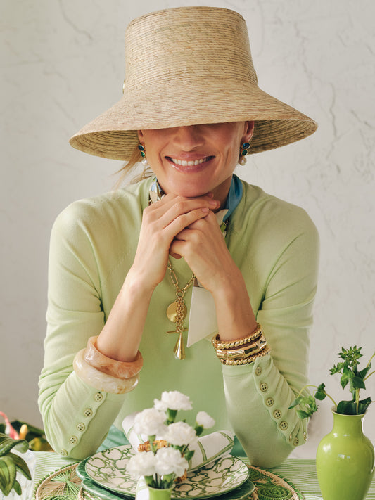 Woman wearing a light green Jamey Sweater by J.McLaughlin paired with layered gold bracelets, statement earrings, and a wide-brim straw hat, embodying effortlessly stylish and enduring designs.