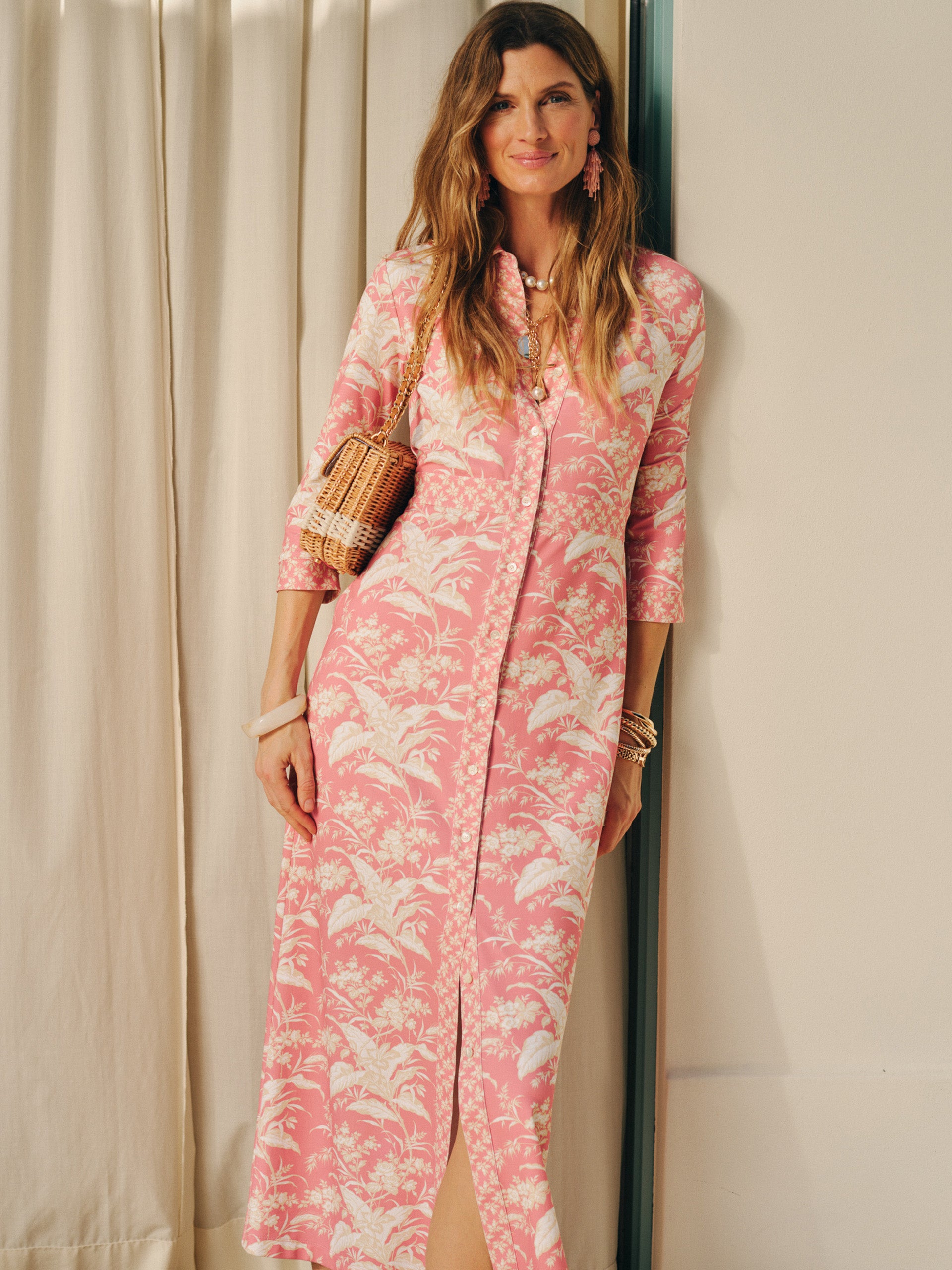 A woman wearing the Kailyn Maxi Dress in Giardino Toile Bud by J.McLaughlin, featuring a soft pink and white botanical print, styled with a woven bag and elegant jewelry, showcasing effortlessly stylish, enduring designs and innovative fabrics. A destination for defining style. Life, Well-Lived.