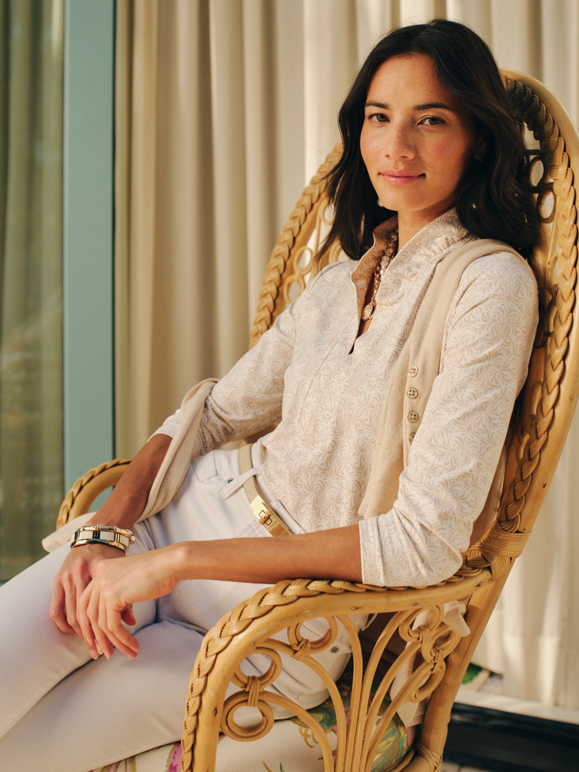 A woman seated in a wicker chair wearing the Kate Ruffle Top in Mini Drawing Room by J.McLaughlin, showcasing effortlessly stylish design and enduring elegance in a softly patterned blouse paired with white pants.