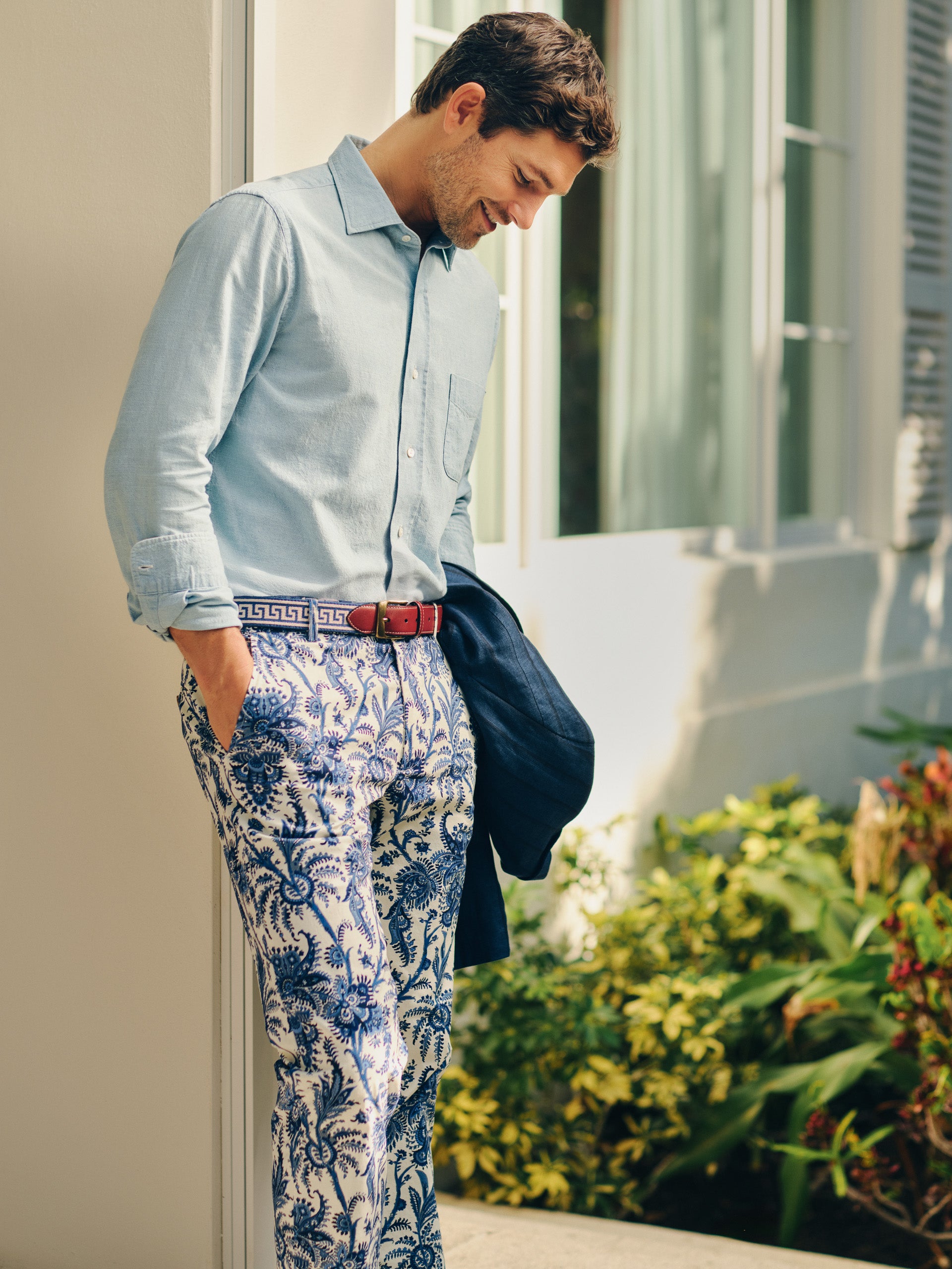 Man wearing Madison Fancy Pants in Heirloom Paisley paired with a light blue shirt and red belt, holding a navy jacket, showcasing J.McLaughlin's effortlessly stylish and enduring designs.