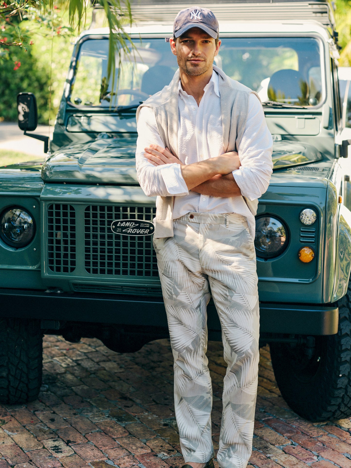 Man wearing Madison Fancy Pants in Vibrant Palms by J.McLaughlin with a white shirt and light sweater draped over shoulders, standing confidently in front of a vintage green Land Rover, showcasing effortlessly stylish and enduring designs. A destination for defining style. Life, Well-Lived.