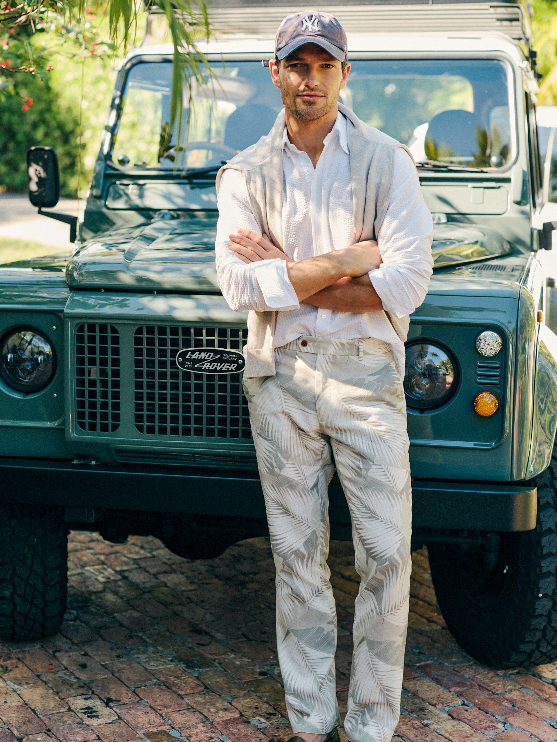 Man wearing Madison Fancy Pants in Vibrant Palms by J.McLaughlin with a white shirt and light sweater draped over shoulders, standing confidently in front of a vintage green Land Rover, showcasing effortlessly stylish and enduring designs. A destination for defining style. Life, Well-Lived.