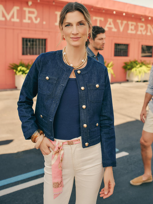 A woman wearing the Jackie Denim Jacket by J.McLaughlin, styled with elegant pearl necklace and light trousers with a printed scarf belt, showcasing effortlessly stylish, enduring designs in a bright outdoor setting, embodying a legacy of timeless fashion and a destination for defining style. Life, Well-Lived.