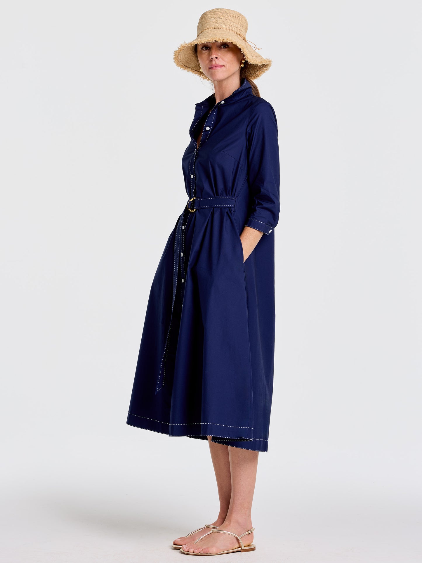 Woman wearing a navy Stephanie Dress by J.McLaughlin with a belted waist, three-quarter sleeves, paired with a woven sun hat and sandals, showcasing effortlessly stylish, enduring designs in innovative fabrics. A destination for defining style. Life, Well-Lived.