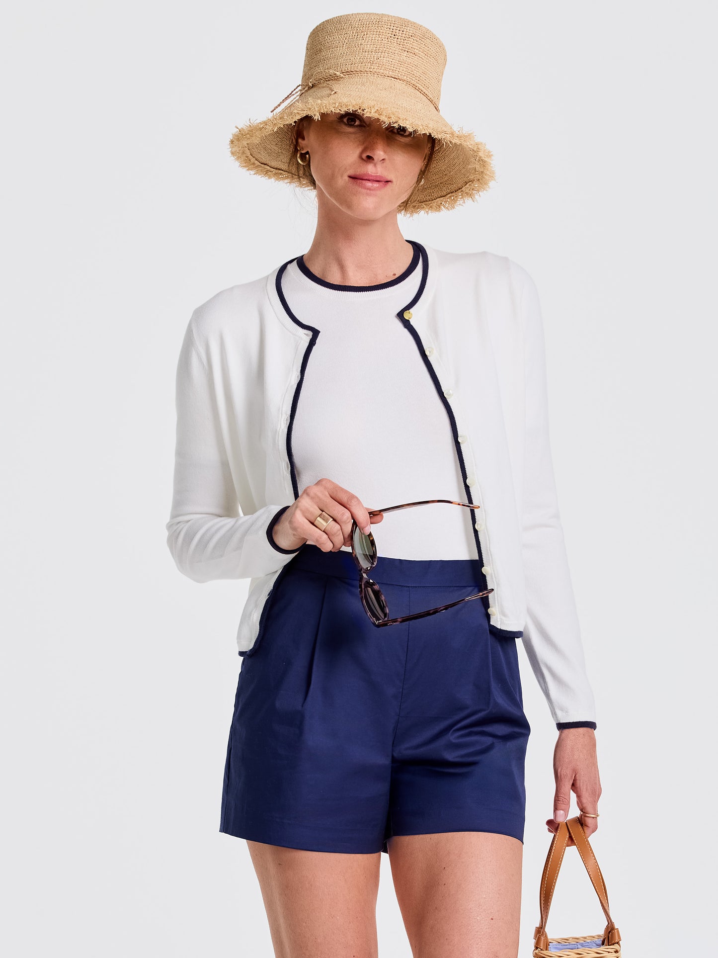 Woman wearing the Aileen Cardigan by J.McLaughlin in white with navy trim, paired with navy shorts and a straw hat, showcasing effortlessly stylish, enduring designs in innovative fabrics.