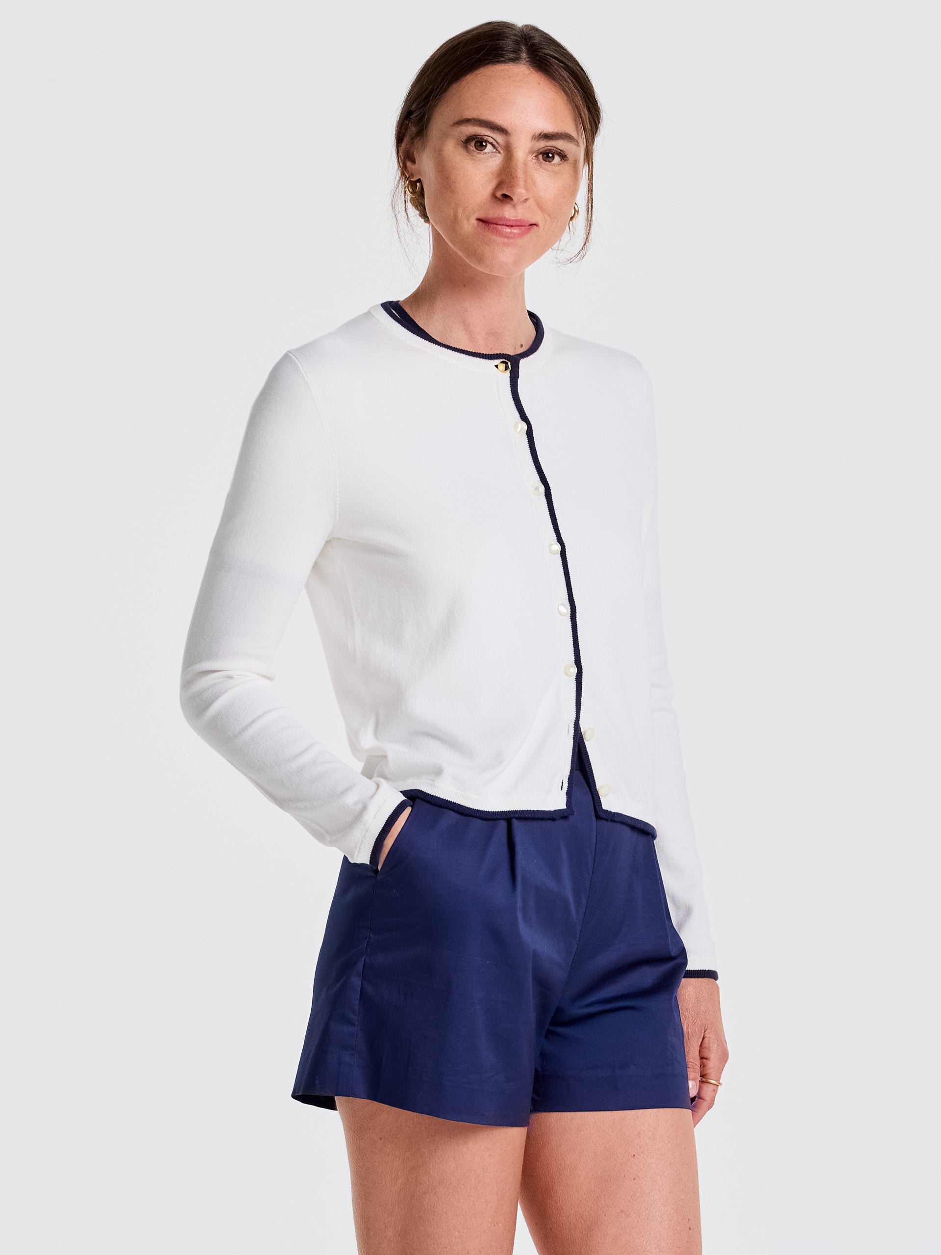 Woman wearing a white Aileen Cardigan with navy trim and pearl buttons paired with navy shorts, showcasing J.McLaughlin’s effortlessly stylish, enduring designs and innovative fabrics.