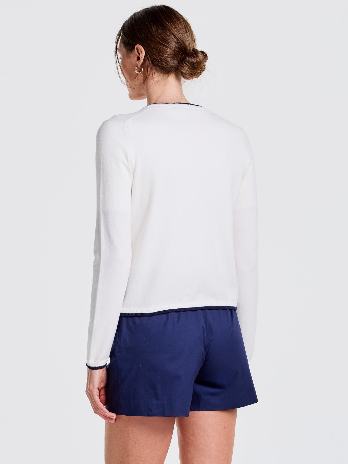 Woman modeling the Aileen Cardigan by J.McLaughlin in white with navy trim, paired with navy shorts, showcasing effortlessly stylish, enduring designs and innovative fabrics.