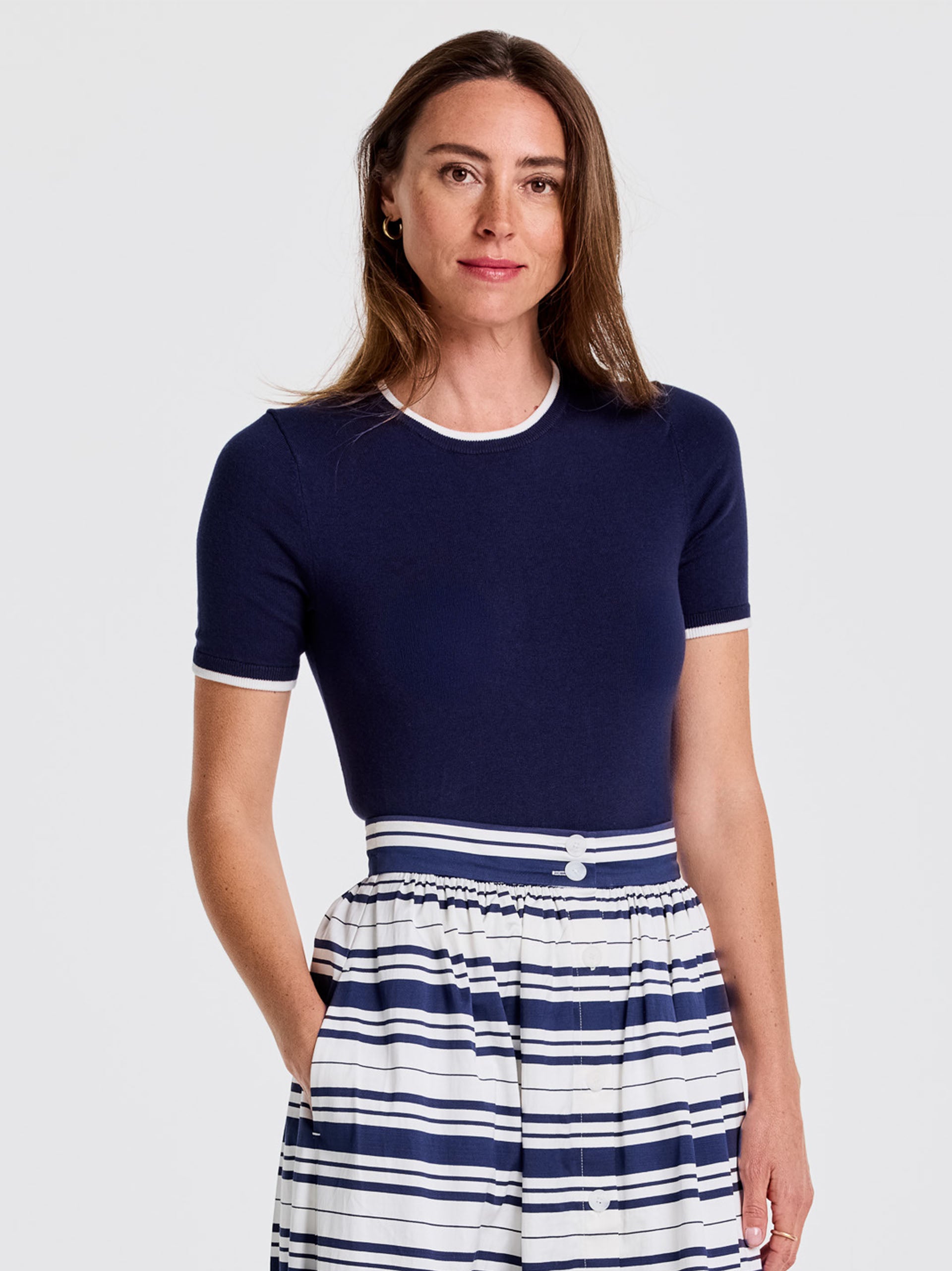 A model wears the Alyson Sweater Tee by J.McLaughlin, a navy short-sleeve sweater with white trim, paired with a striped skirt, showcasing effortlessly stylish, enduring designs and innovative fabrics from a destination for defining style. Life, Well-Lived.