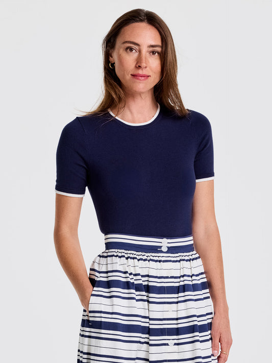 A model wears the Alyson Sweater Tee by J.McLaughlin, a navy short-sleeve sweater with white trim, paired with a striped skirt, showcasing effortlessly stylish, enduring designs and innovative fabrics from a destination for defining style. Life, Well-Lived.
