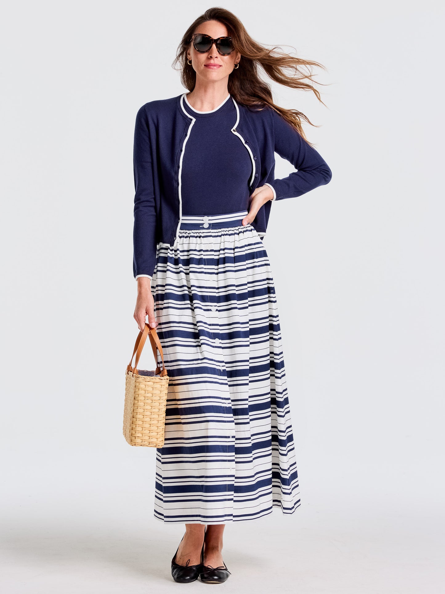 Woman wearing J.McLaughlin Susie Skirt in Mariner Stripe with navy top and cardigan, carrying a woven bag, epitomizing effortlessly stylish looks with enduring designs and timeless fashion.