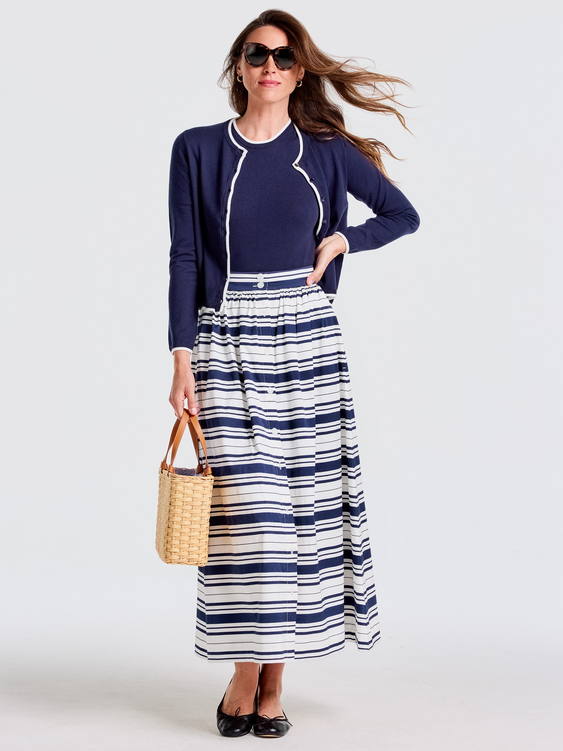 Woman wearing J.McLaughlin Susie Skirt in Mariner Stripe with navy top and cardigan, carrying a woven bag, epitomizing effortlessly stylish looks with enduring designs and timeless fashion.