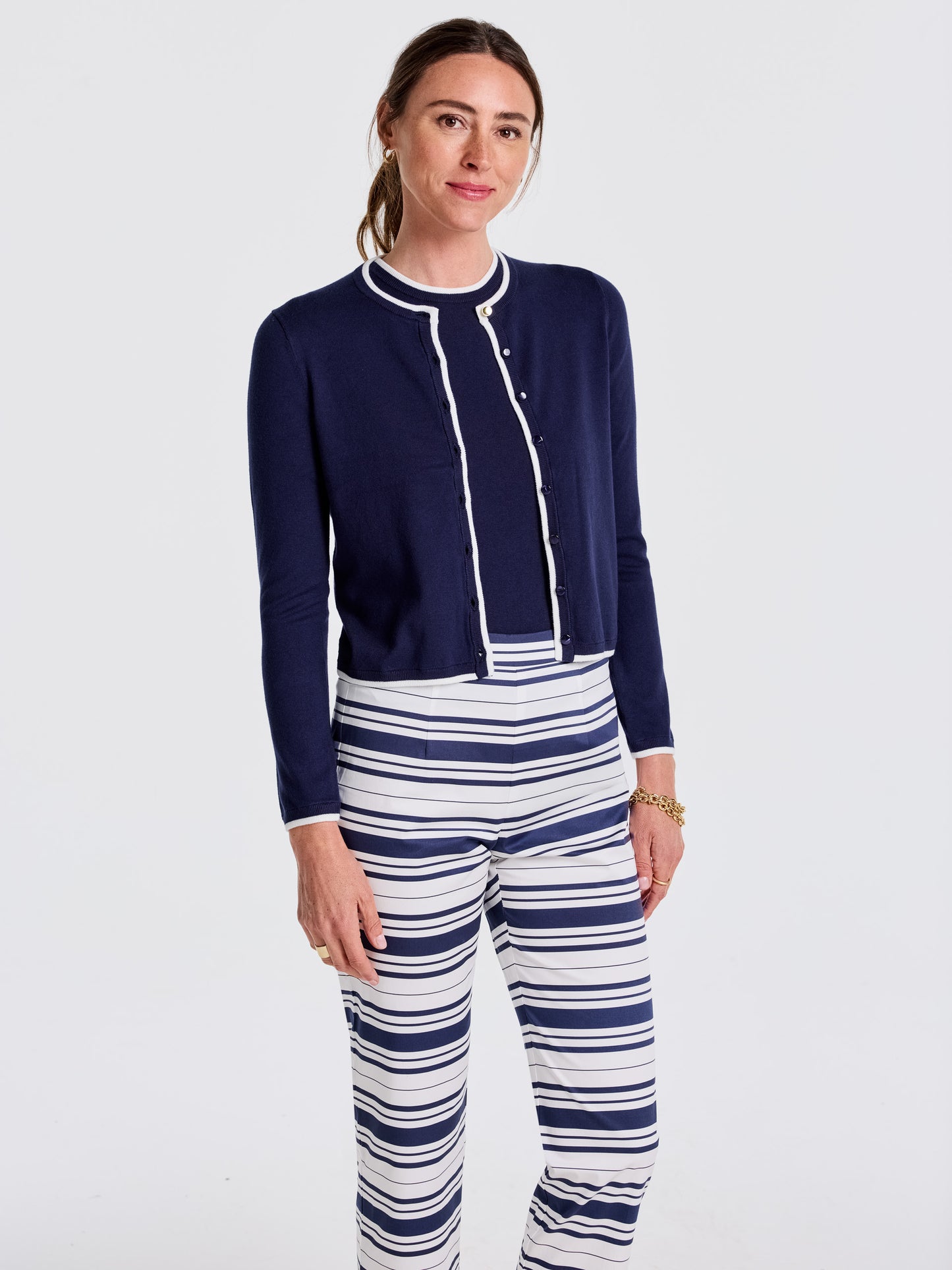 Woman wearing an effortlessly stylish Aileen Cardigan by J.McLaughlin in navy with white trim, paired with striped pants, showcasing enduring designs and innovative fabrics.