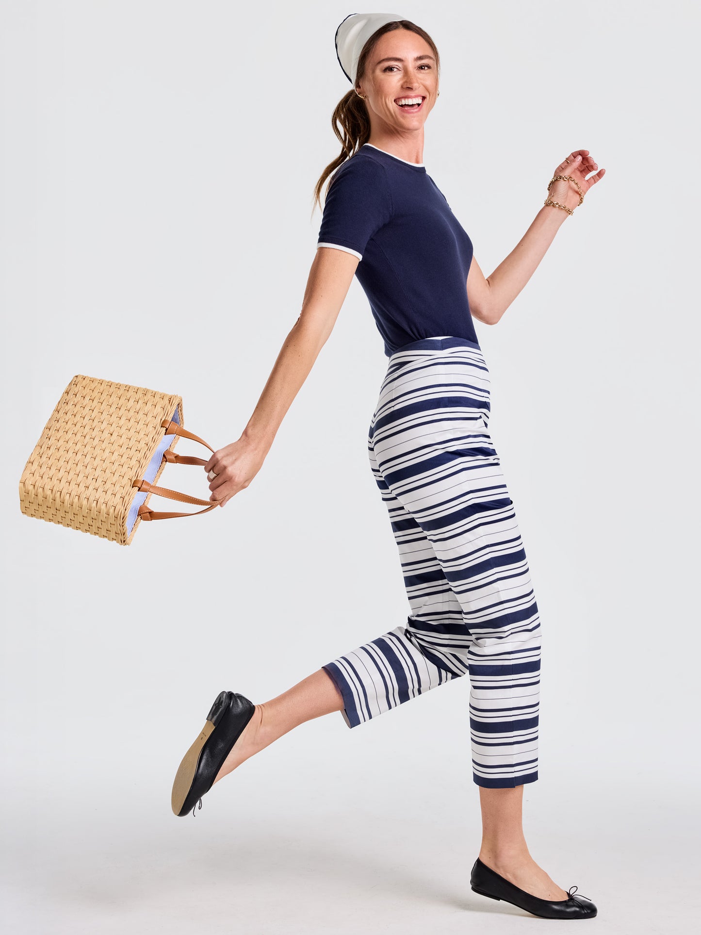 A joyful woman wearing J.McLaughlin Betina Pants in Mariner Stripe paired with a navy top, black flats, and carrying a woven tote, embodying effortlessly stylish, enduring designs.