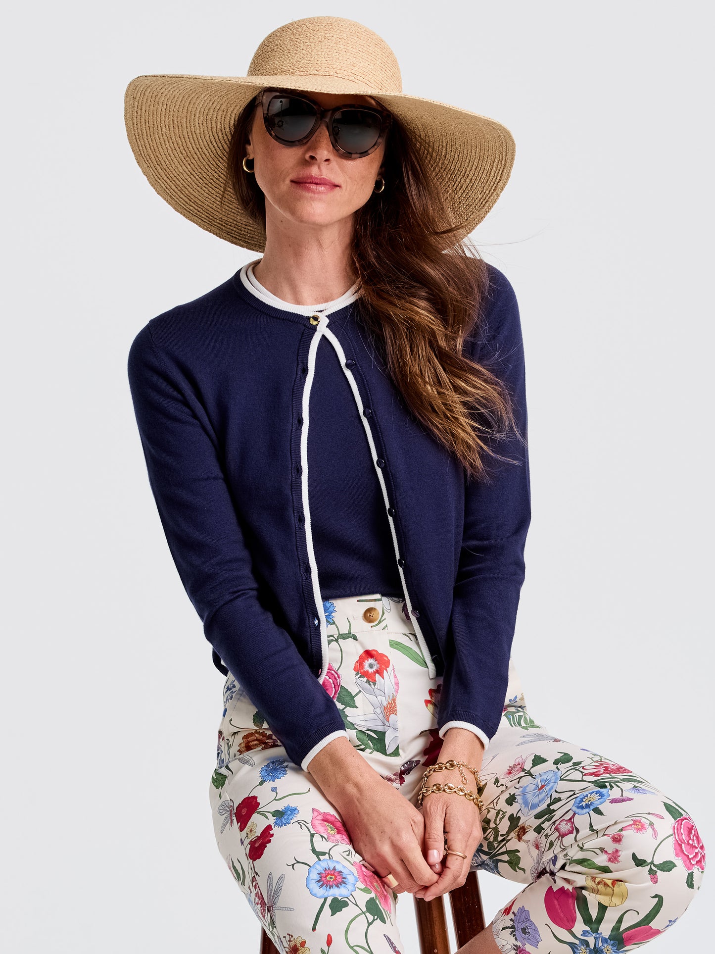 Woman wearing a navy Aileen Cardigan by J.McLaughlin, styled with floral pants, oversized hat, and sunglasses, showcasing effortlessly stylish, enduring designs and innovative fabrics.