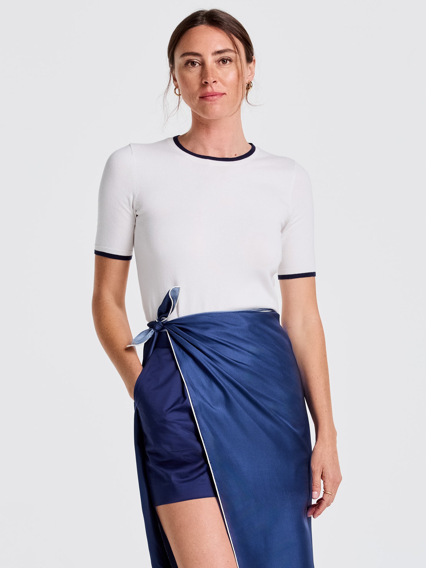 A woman modeling the Alyson Sweater Tee by J.McLaughlin, a white short-sleeve sweater tee with navy trim at collar and sleeves, paired with a silky navy wrap skirt, showcasing effortlessly stylish, enduring designs and innovative fabrics for a life well-lived.