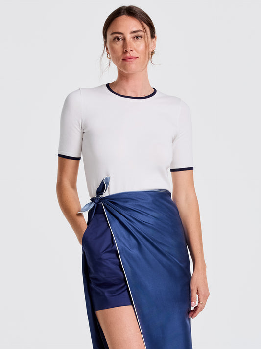 A woman modeling the Alyson Sweater Tee by J.McLaughlin, a white short-sleeve sweater tee with navy trim at collar and sleeves, paired with a silky navy wrap skirt, showcasing effortlessly stylish, enduring designs and innovative fabrics for a life well-lived.