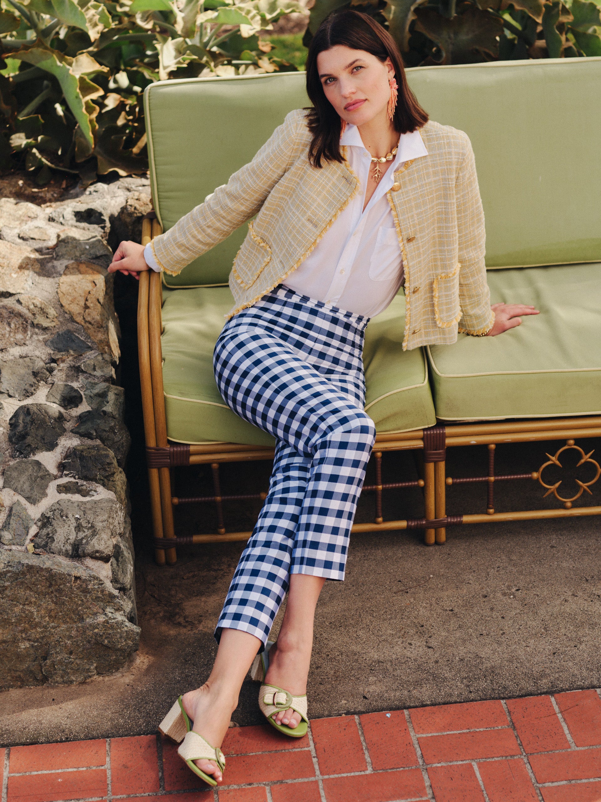 A woman lounges on a green cushioned bench wearing Millbrook Silk Blend Pants in Gingham by J.McLaughlin, paired with a classic yellow textured jacket and white blouse, embodying effortlessly stylish, enduring designs and innovative fabrics from a destination for defining style. Life, Well-Lived.