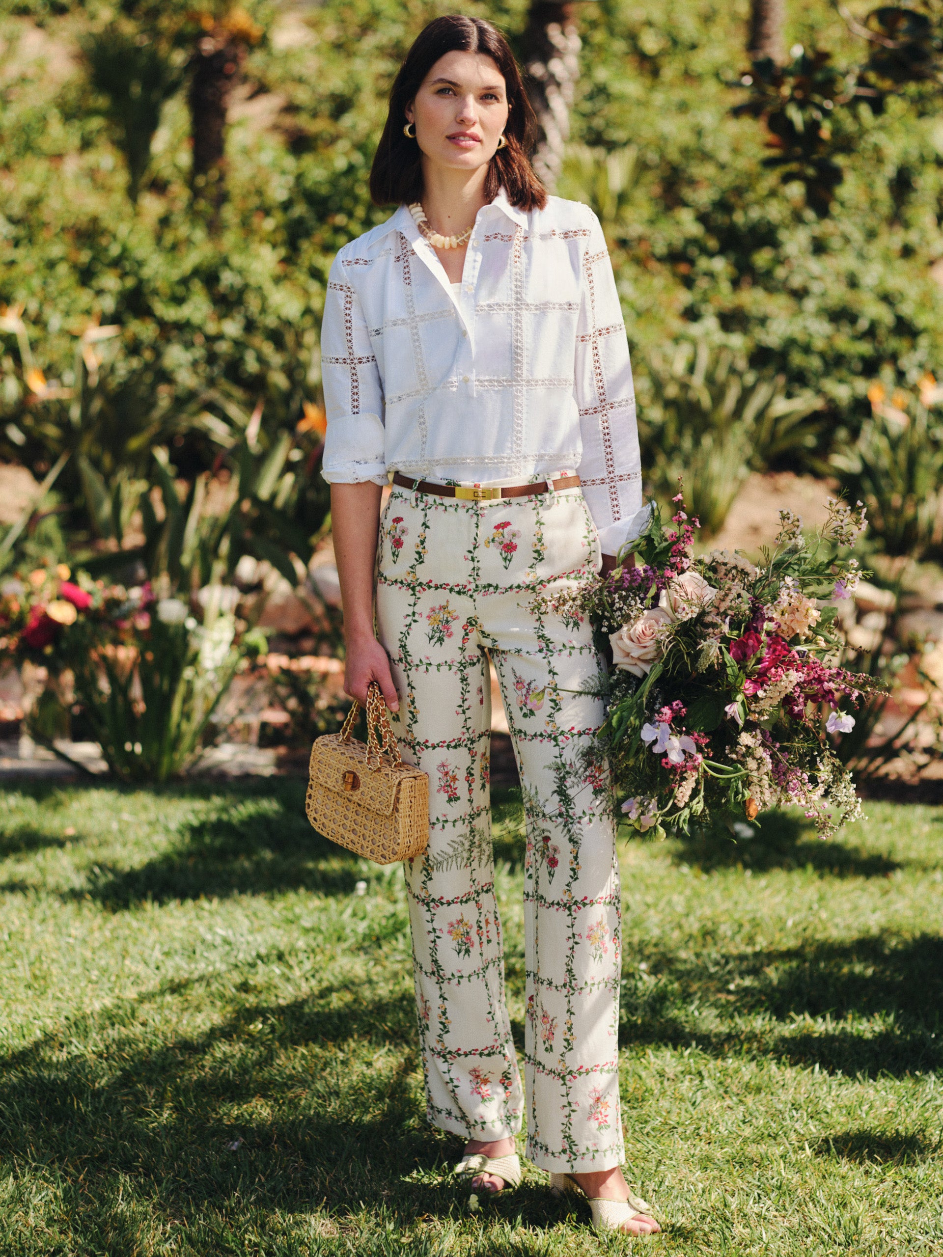 Model wearing J.McLaughlin Helen Linen Blend Pants in Floral Medley Frame paired with a white blouse, holding a woven handbag and a bouquet of flowers in a garden setting, showcasing effortlessly stylish, enduring designs with innovative fabrics for a life well-lived.